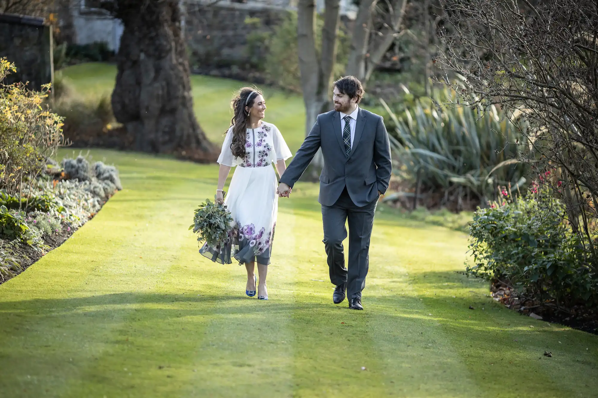 A couple walk hand in hand on a green lawn surrounded by trees and plants. The woman wears a white dress with floral designs and holds a bouquet, while the man wears a dark suit. They smile at each other in a sunny, peaceful garden setting.
