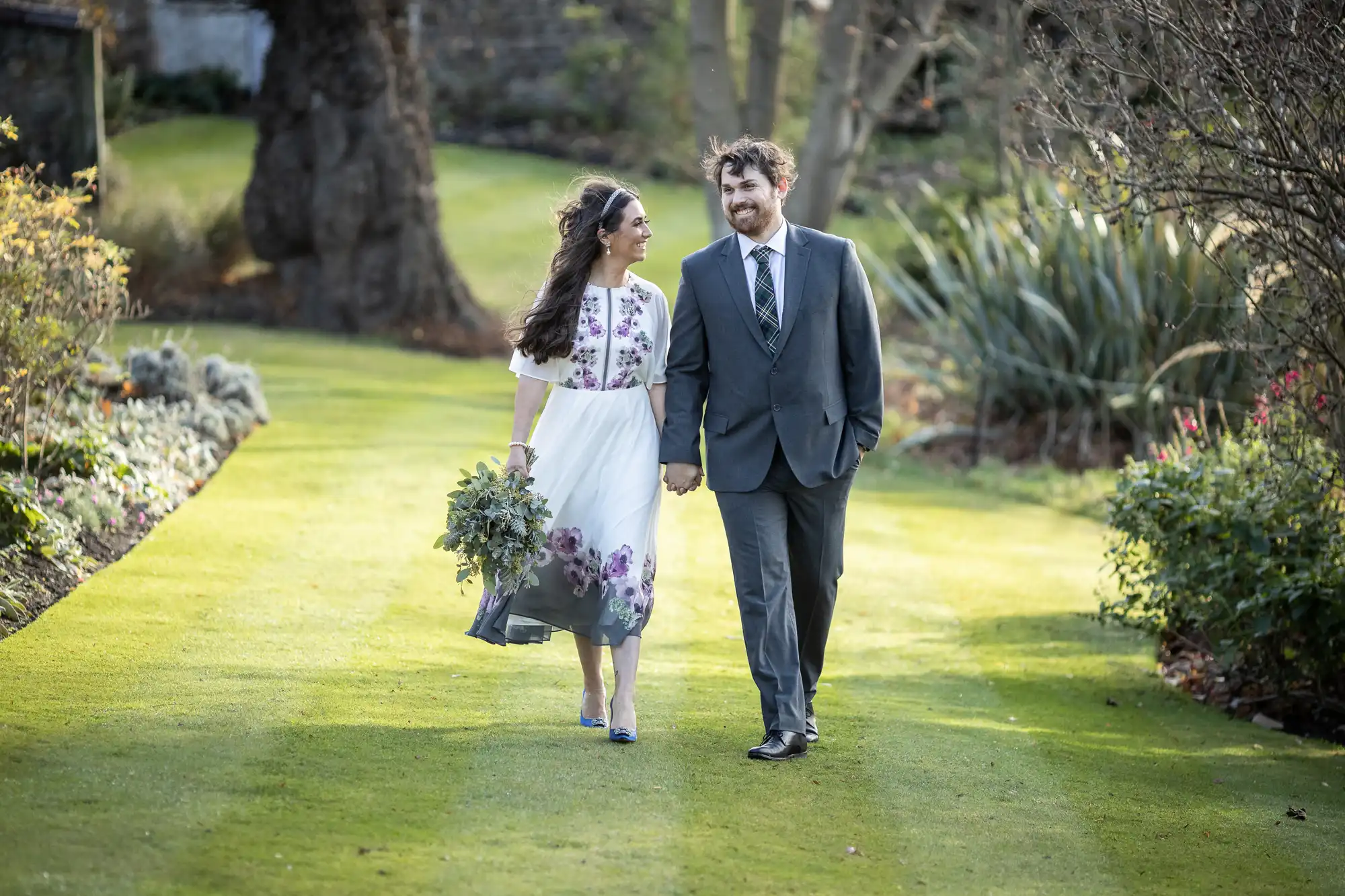 A couple walks hand-in-hand on a green lawn. The woman wears a white floral dress and holds a bouquet, while the man wears a suit and tie. They smile at each other, surrounded by trees and garden plants in a sunlit outdoor setting.