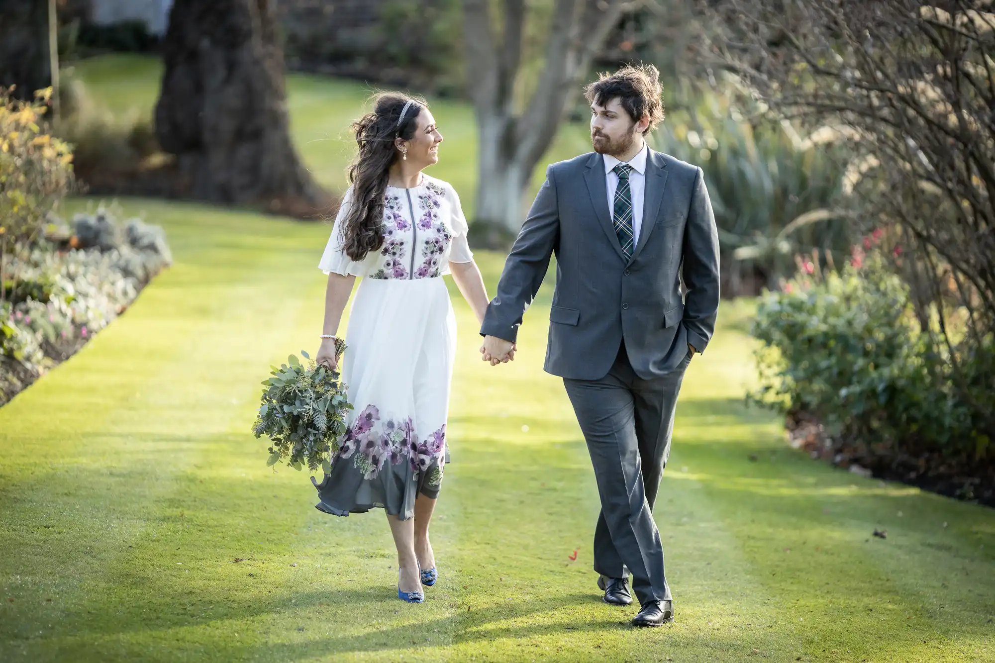 A woman in a white dress with purple floral patterns holds greenery and walks hand in hand with a man in a grey suit and checked tie. They are smiling at each other whilst walking on a sunlit lawn with trees and gardens in the background.