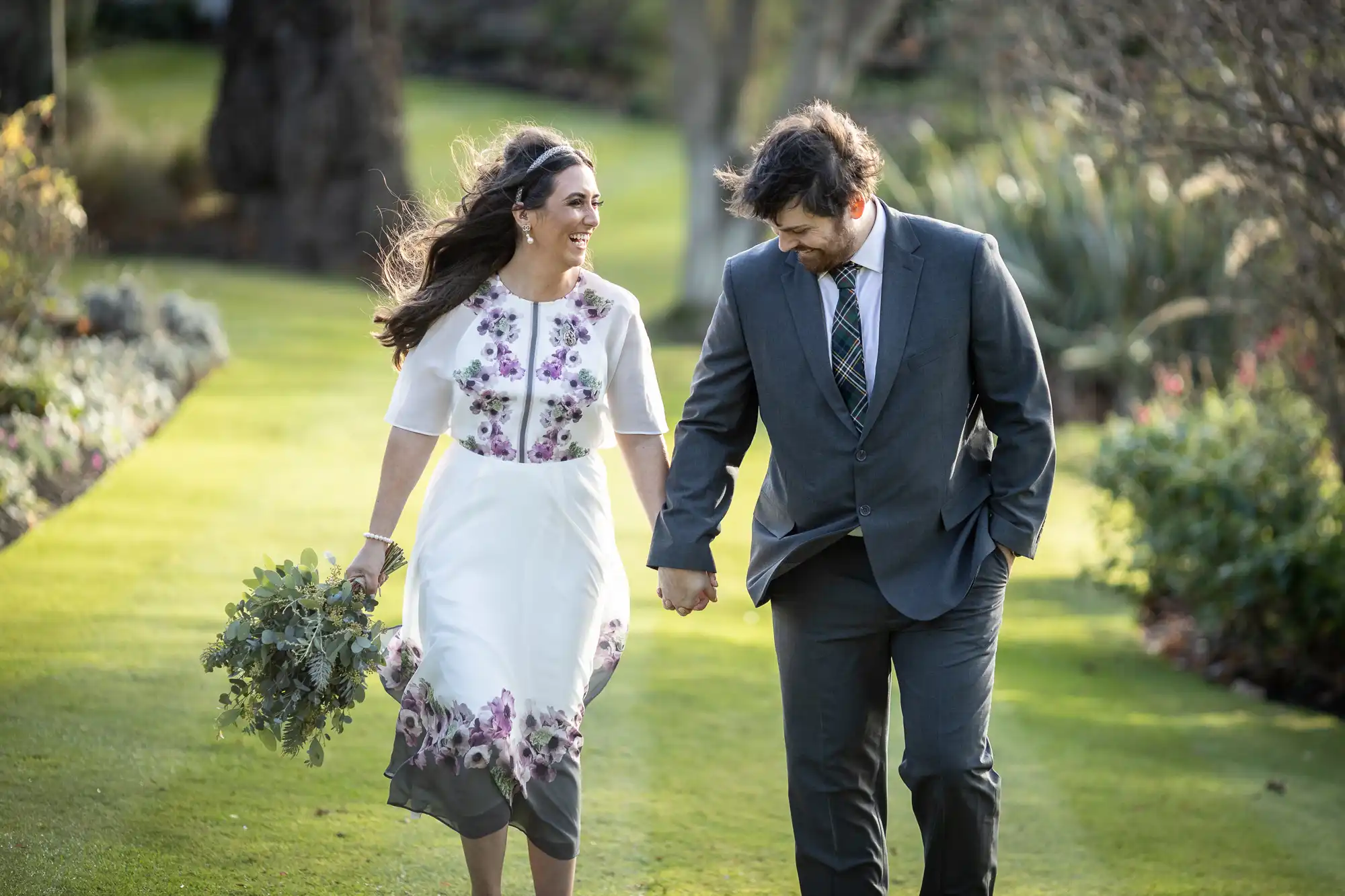A woman in a white dress with purple floral patterns holds a bouquet and walks hand-in-hand with a man in a grey suit and checked tie. They smile at each other whilst walking on a sunlit garden path surrounded by greenery.