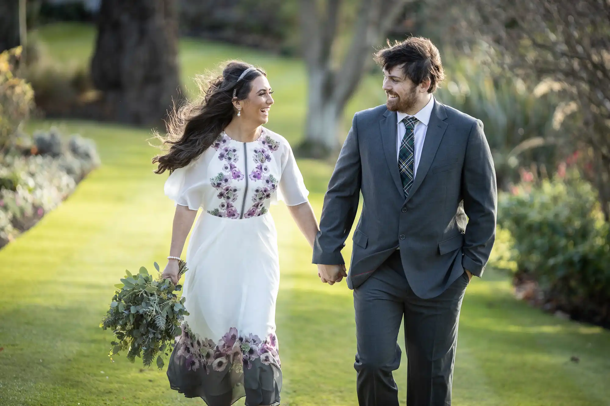 A smiling couple hold hands while walking outdoors on a sunny day. The woman wears a white dress with floral patterns and carries a bouquet. The man wears a grey suit and checked tie. They look at each other, walking on a lawn with trees and plants in the background.