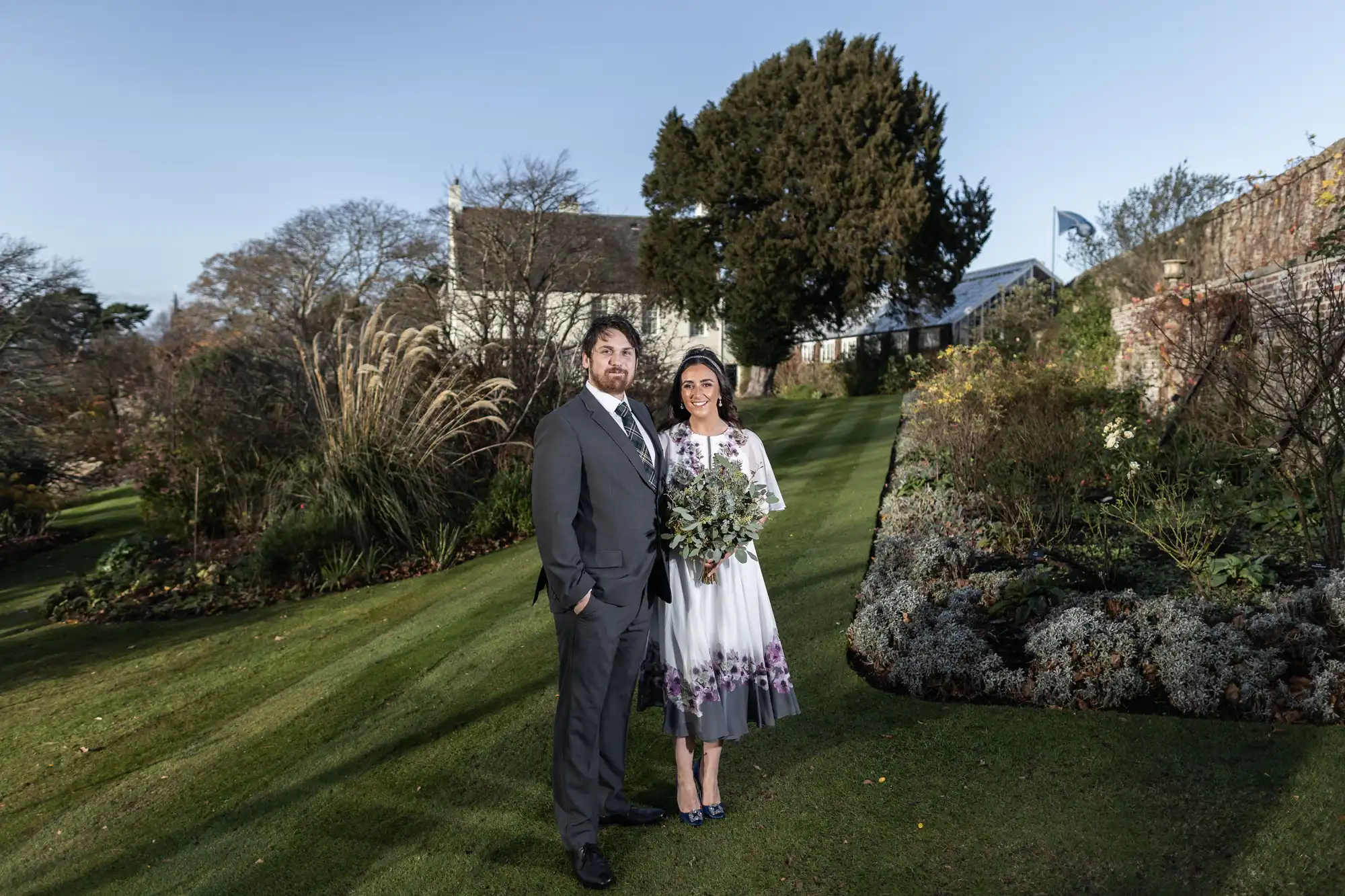 A couple pose on a manicured lawn in front of a landscaped garden. The man wears a grey suit, and the woman wears a white dress with purple floral details, holding a bouquet. Behind them are trees, shrubs, and a large house with a sloping roof.