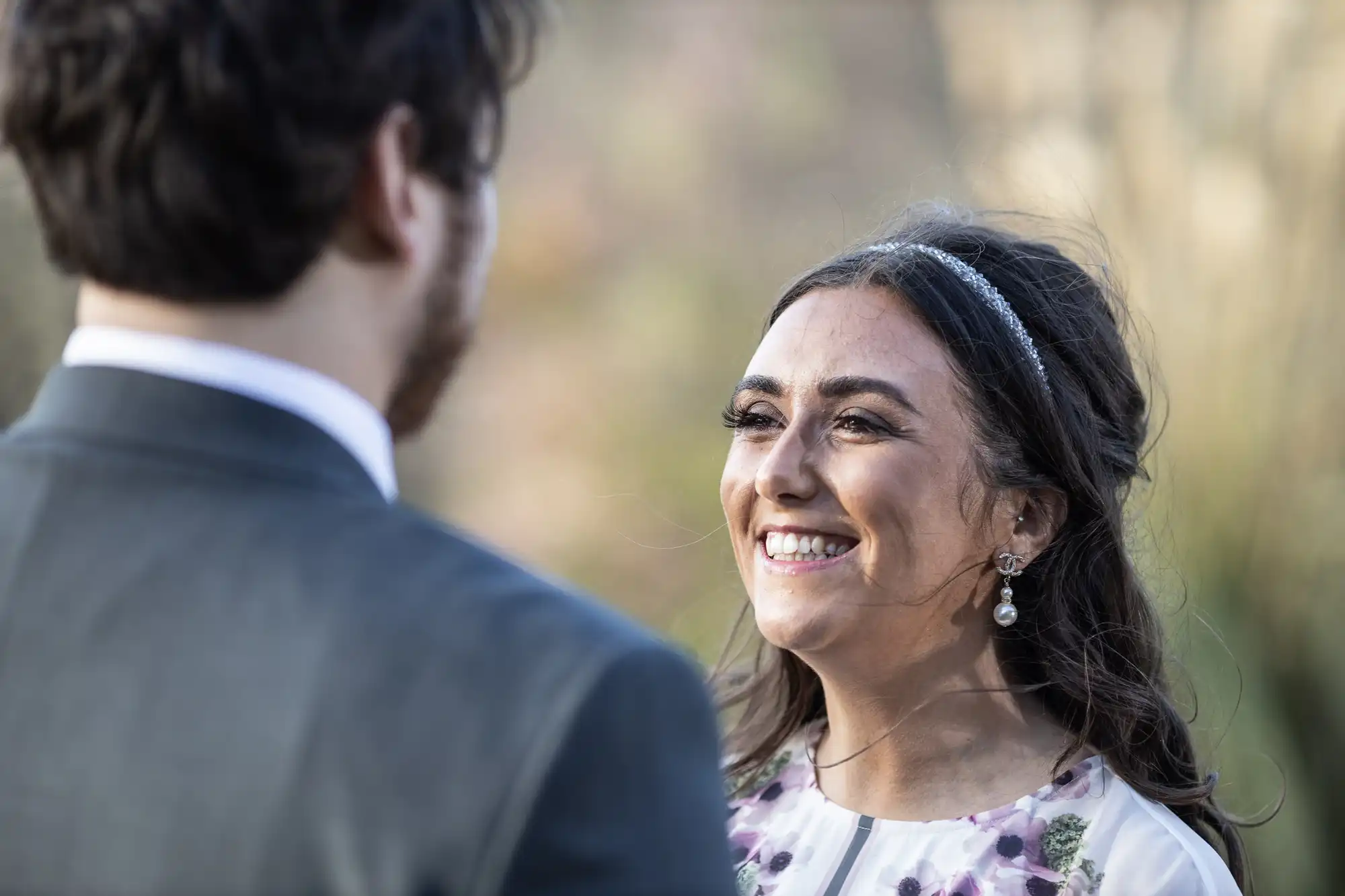 A smiling woman with long dark hair, wearing floral earrings and a headband, faces a man in a suit. The background is softly blurred, suggesting an outdoor setting with natural light. The woman appears joyful and engaged in conversation.