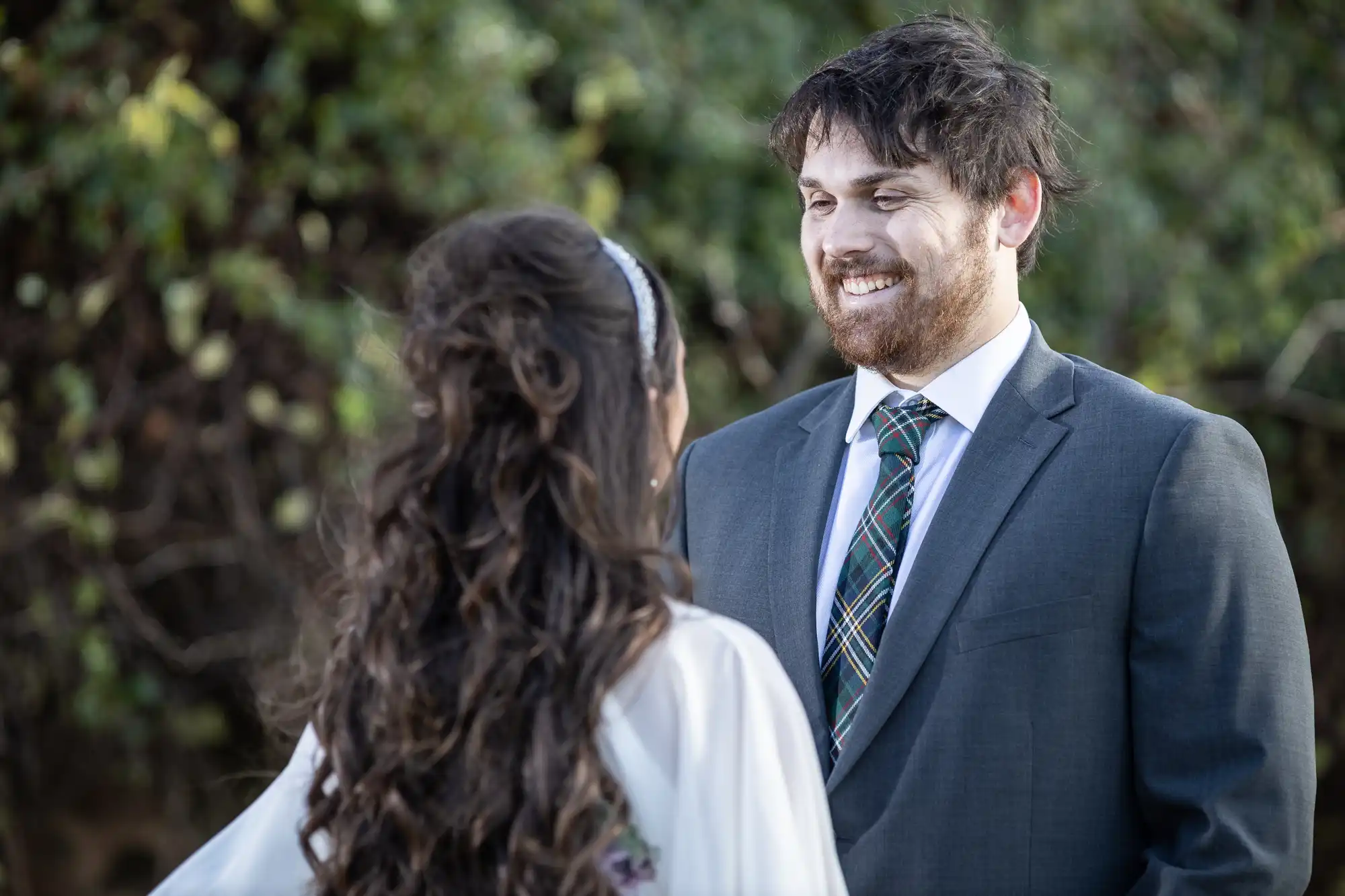 A man in a suit and checked tie smiles warmly at a woman with long brown wavy hair facing him. They stand outdoors with green foliage in the blurred background, sharing an intimate, joyful moment.