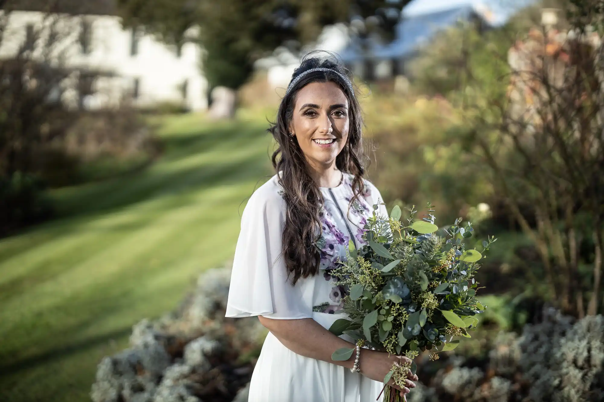 A woman in a white dress stands outdoors on a grassy lawn, holding a bouquet of green and white flowers. She has long, wavy brown hair and is smiling at the camera. Bushes and trees fill the background, with a white house visible in the distance.
