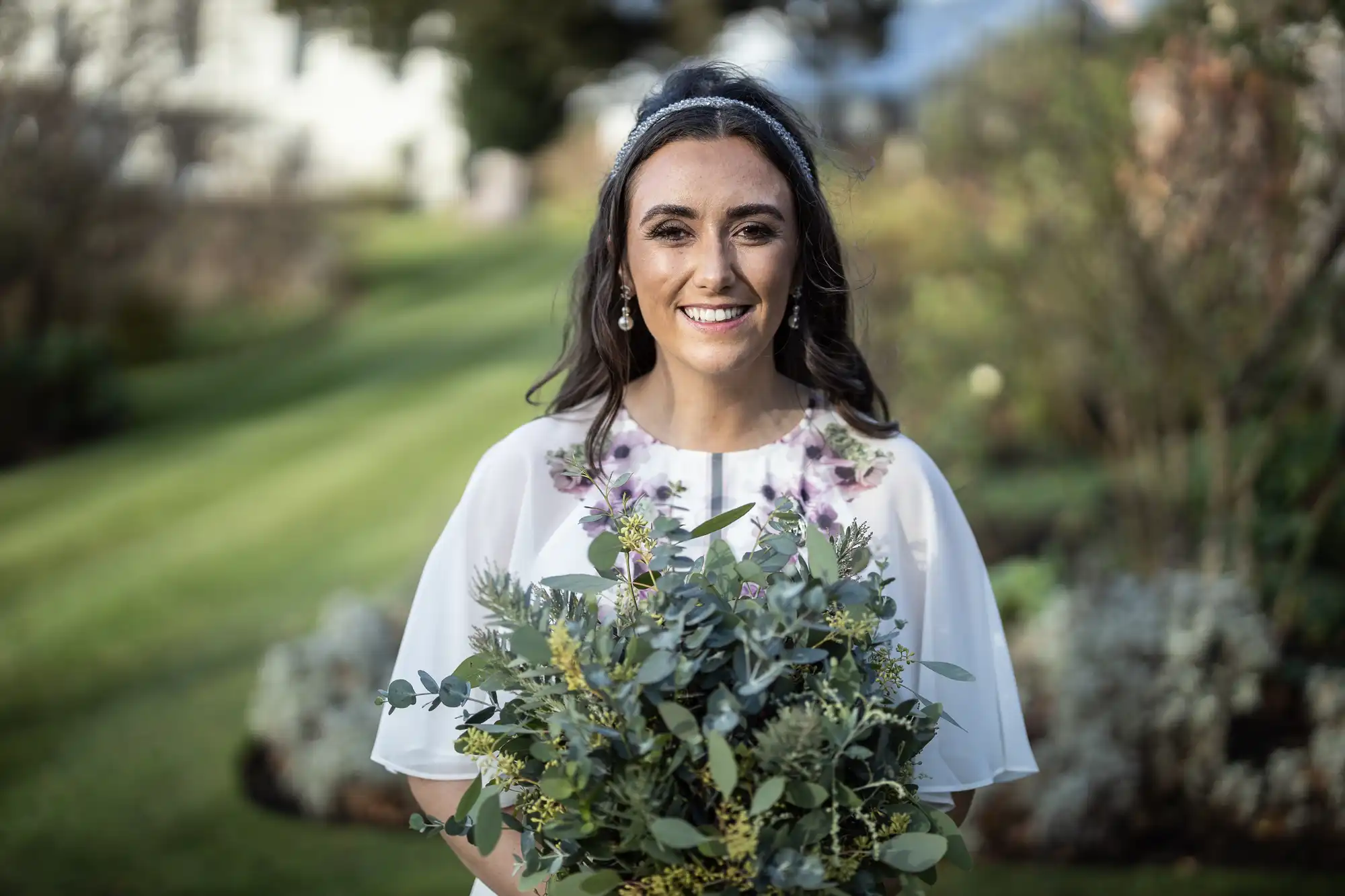 A smiling woman with long dark hair, wearing a white dress with floral details and a headband, stands outdoors holding a large green bouquet. The blurred background features grass, shrubs, and trees, suggesting a garden setting.