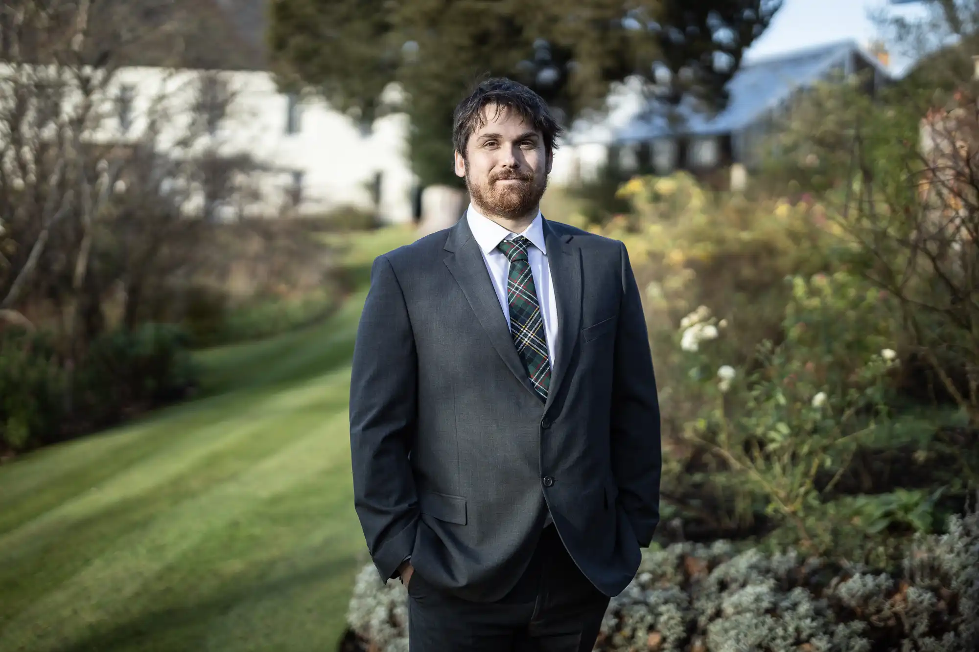 A man in a dark suit and checked tie stands outdoors with his hands in his pockets. He has dark hair and a beard, and is looking at the camera, standing on a manicured lawn with greenery, flowers, and a white building in the background.