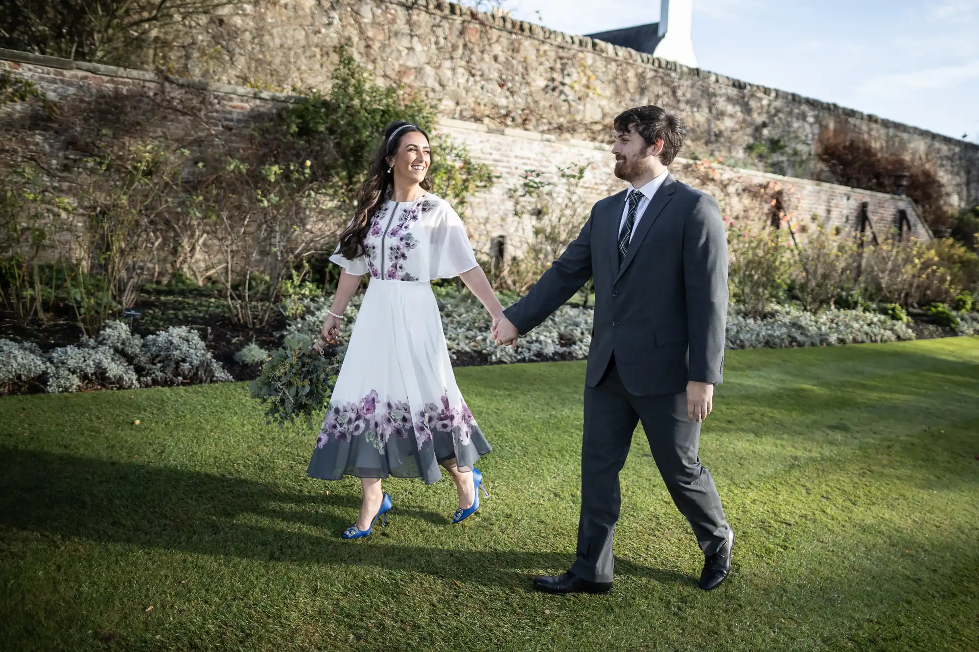 A smiling couple walk hand-in-hand on green grass beside a stone wall. The woman wears a white dress with purple floral designs and blue shoes, while the man is in a grey suit and tie. They look at each other, appearing happy and relaxed outdoors.