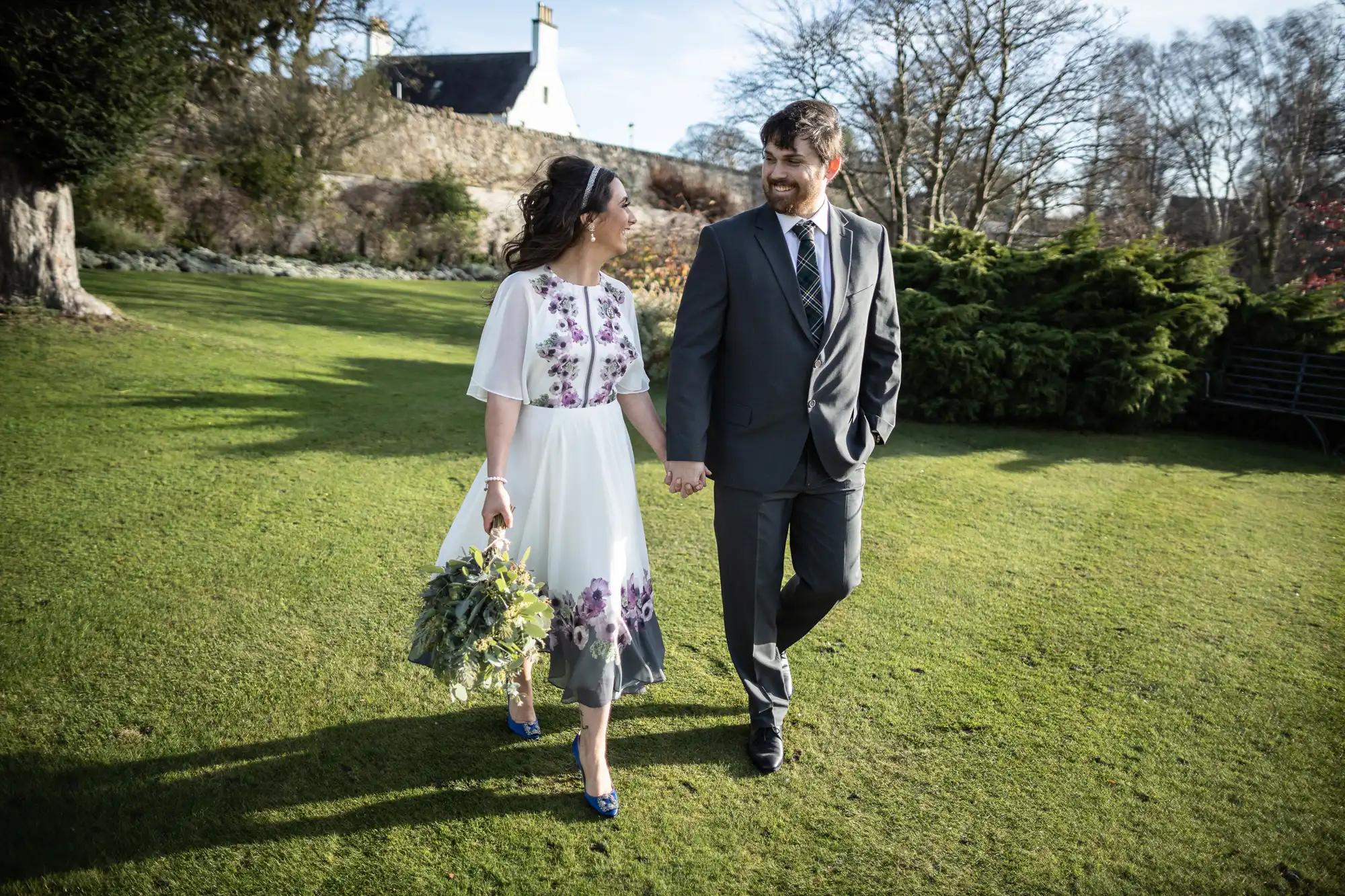 A couple walk hand-in-hand on grass. The woman wears a white dress with purple flowers and blue shoes, holding a bouquet. The man wears a grey suit and tie. Both smile at each other. Trees, hedges, and a house are in the sunlit background.