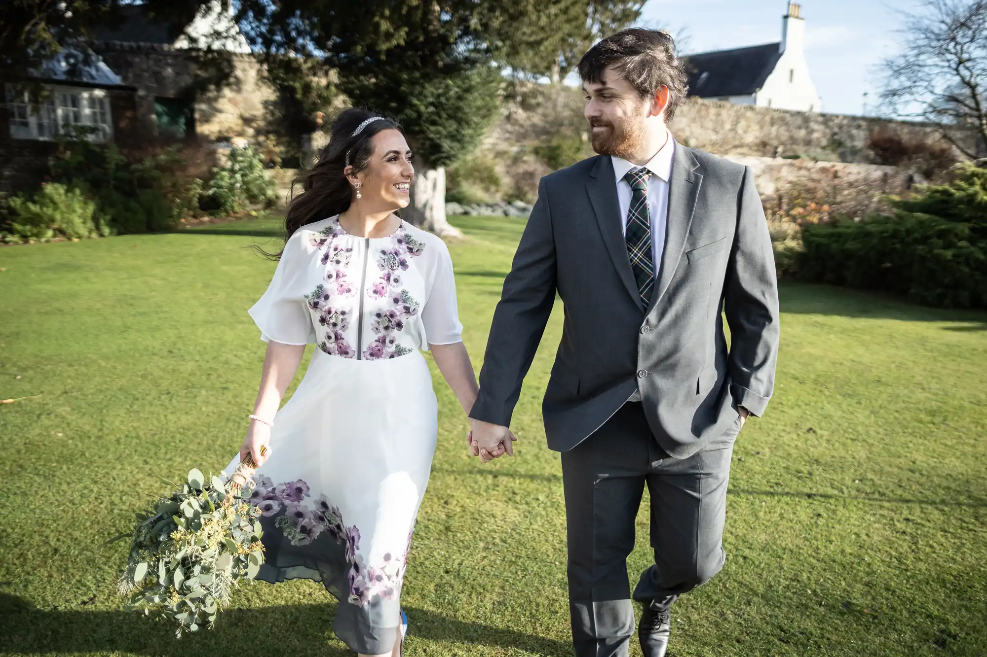A smiling couple hold hands, walking on grass. The woman wears a white dress with purple floral patterns and holds a bouquet; the man wears a grey suit with a checked tie. They look at each other lovingly with a cottage and trees in the background.