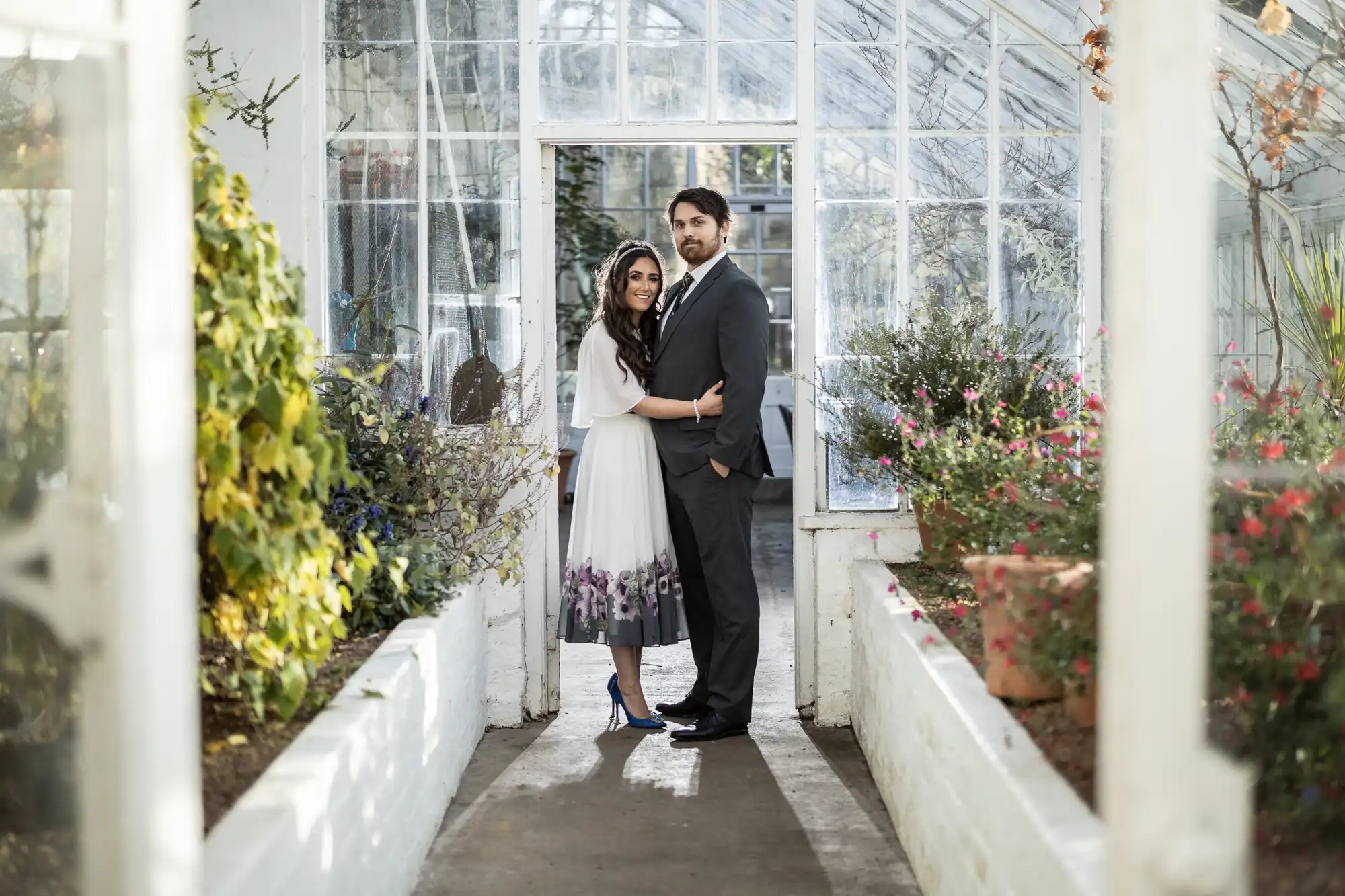A couple stands close together in a glasshouse walkway, surrounded by lush green plants. The woman wears a white dress with purple flowers at the hem and blue heels. The man wears a grey suit and black shoes. Sunlight streams through the glass walls behind them.