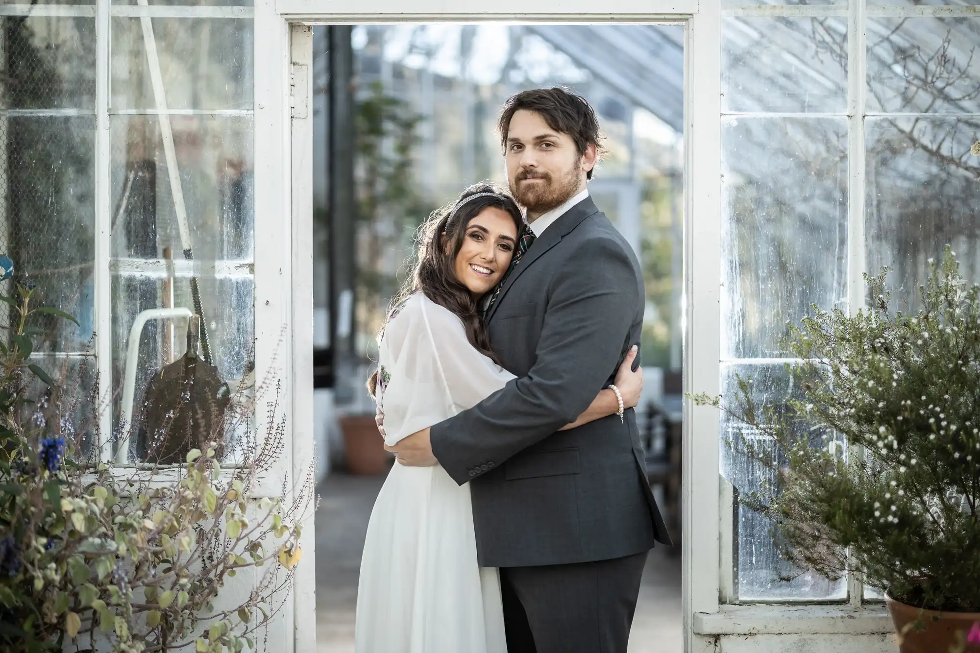 A woman in a white dress lovingly hugs a man in a grey suit inside a glasshouse. They stand framed by open doors, smiling gently at the camera, surrounded by green plants and soft natural light.