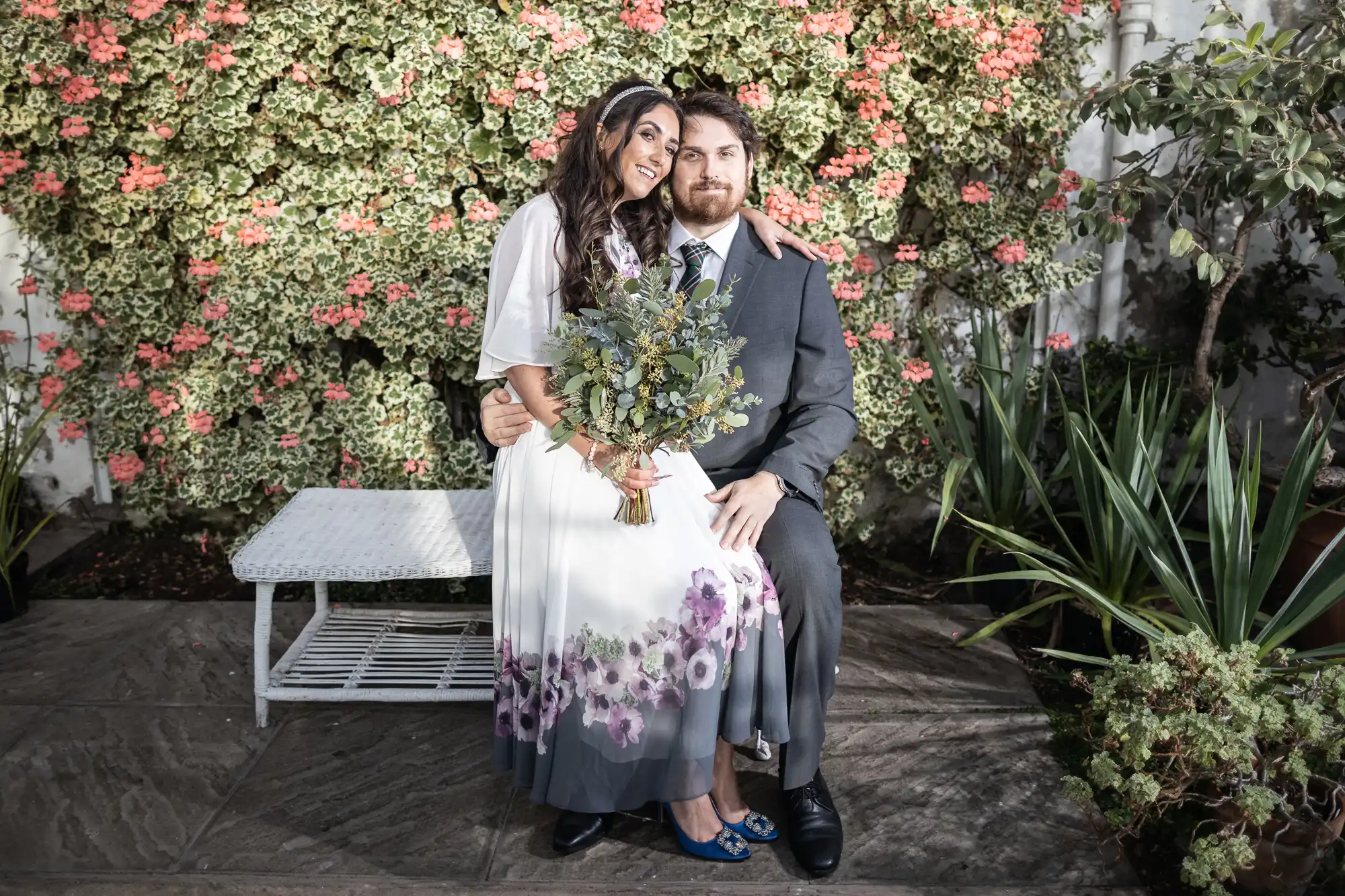 A woman in a white dress with purple flower patterns holds a bouquet and stands close to a man in a grey suit who is seated on a white bench. They are surrounded by lush green and pink flowering plants in an outdoor garden setting.