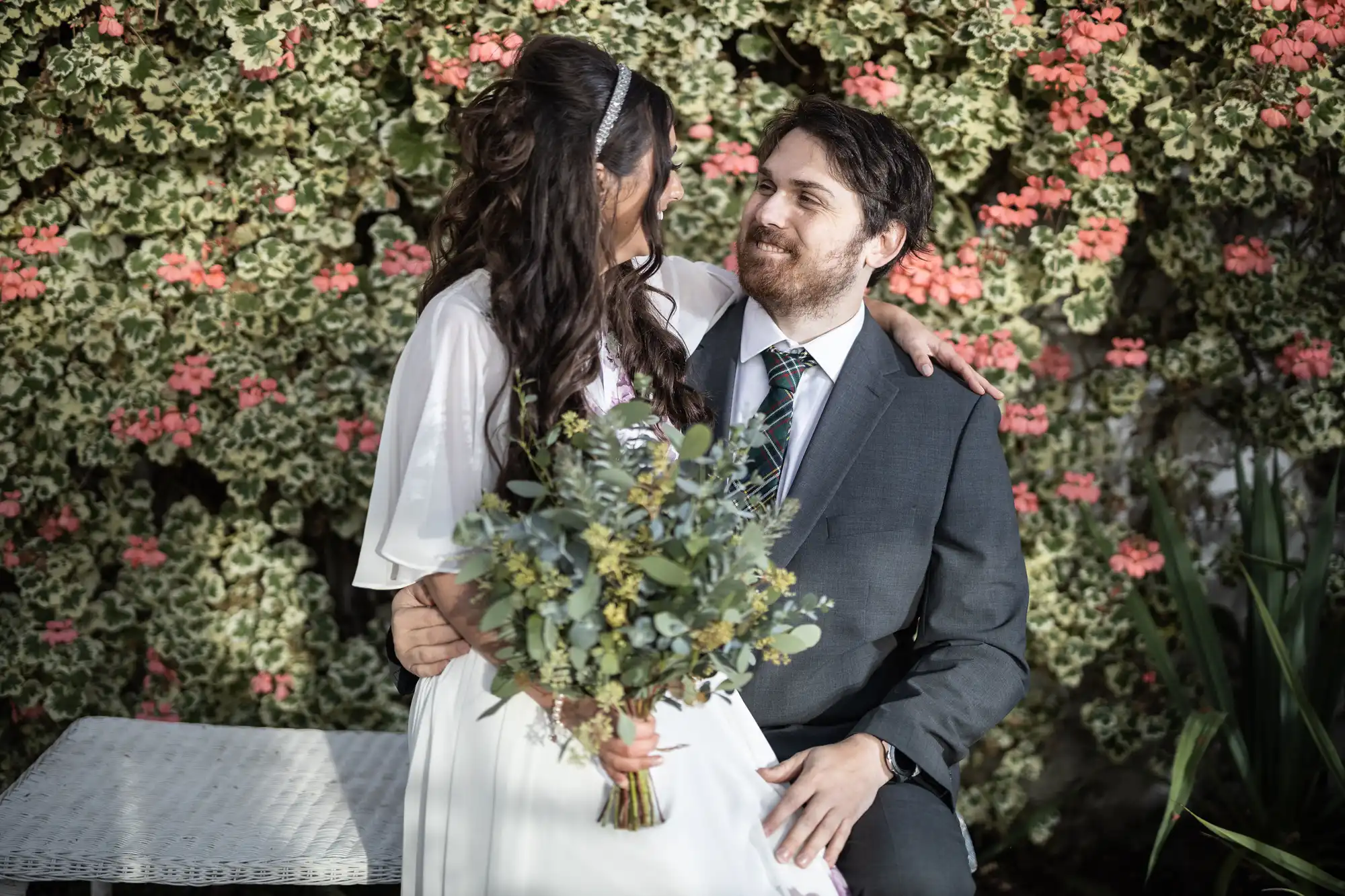 A bride in a white dress holding a bouquet sits on the lap of a groom in a suit. They gaze at each other and smile, surrounded by green foliage with clusters of pink flowers, outdoors on a sunny day.