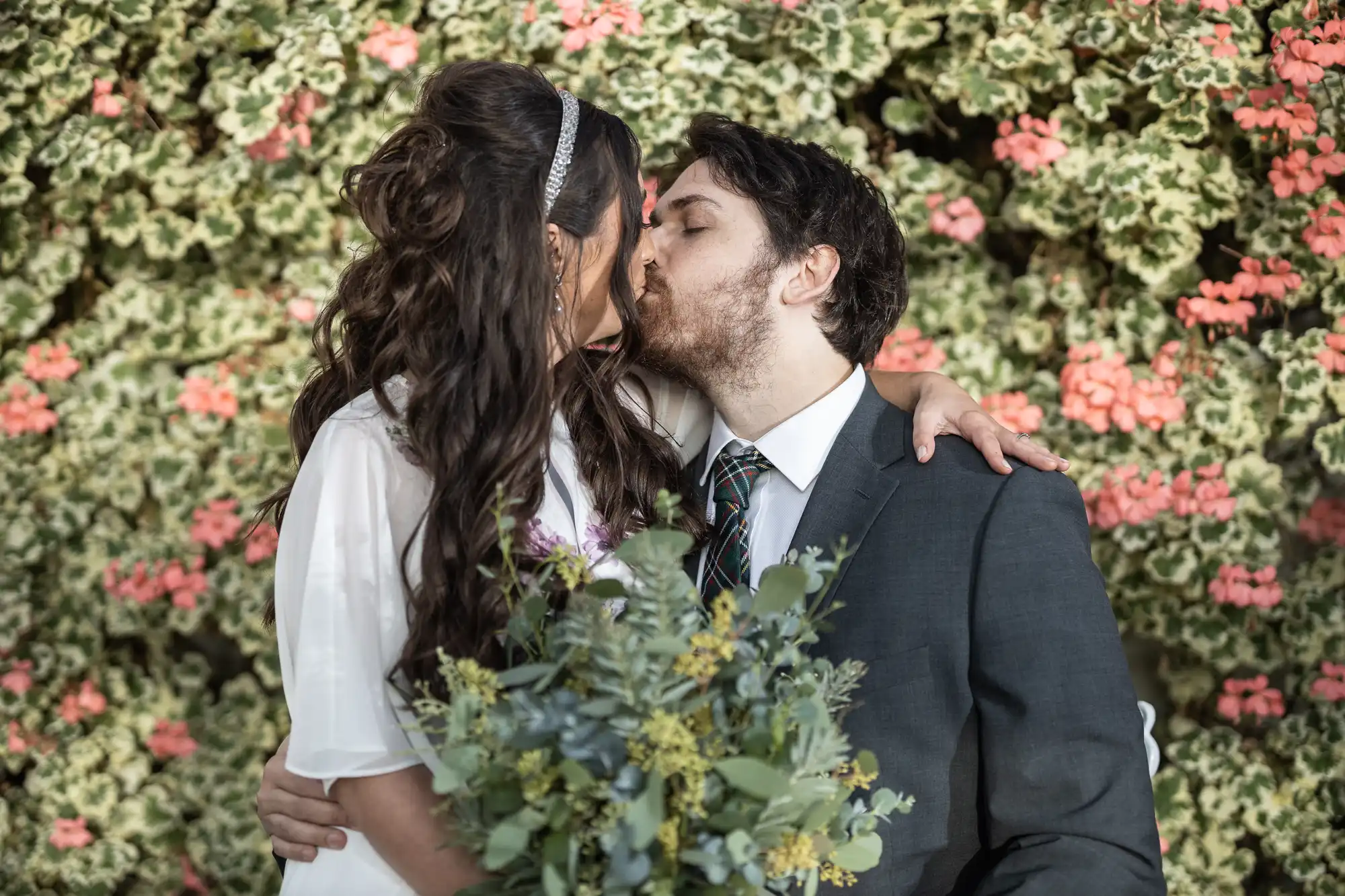 A couple snog in front of a wall covered with green leaves and pink flowers. The woman, in a white dress, holds a bouquet of greenery and has her arm round the man, who is wearing a dark suit and tie. Both have dark hair and look affectionate.