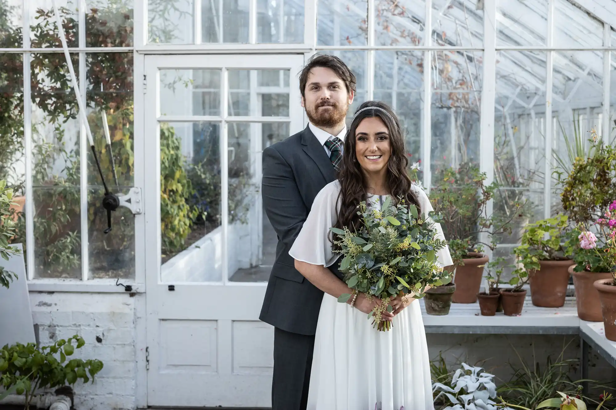 A man in a suit stands behind a woman in a white dress holding a bouquet of greenery. They both smile, posing inside a bright glasshouse with glass walls, surrounded by potted plants and flowers on a white bench.