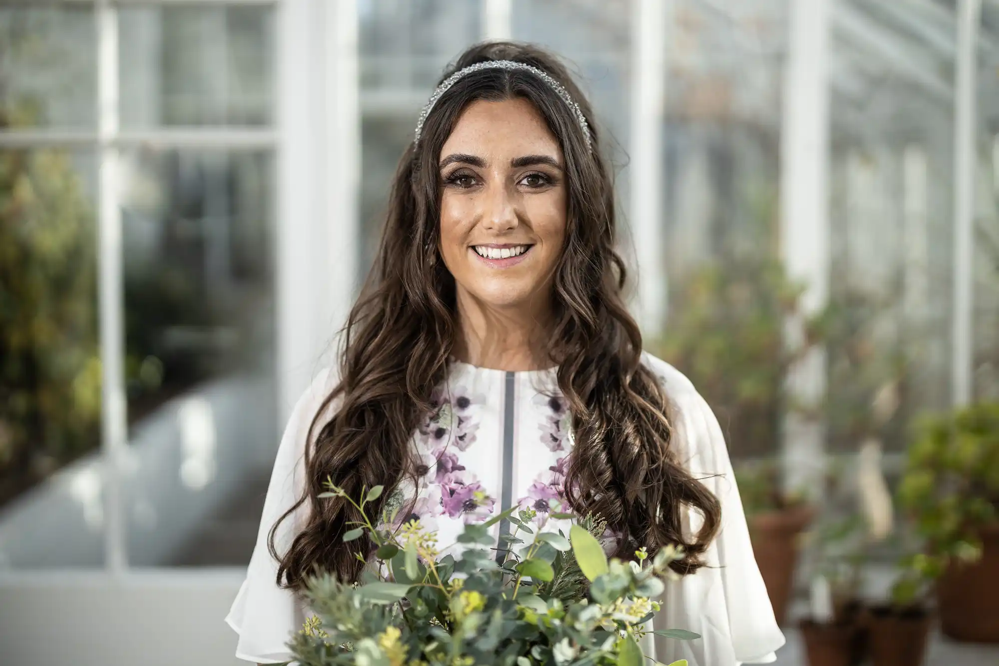 A smiling woman with long, wavy dark hair wears a white dress with purple floral patterns and a headband. She holds a bouquet of green foliage and small flowers. She stands indoors, with bright, blurred glass windows and plants in the background.