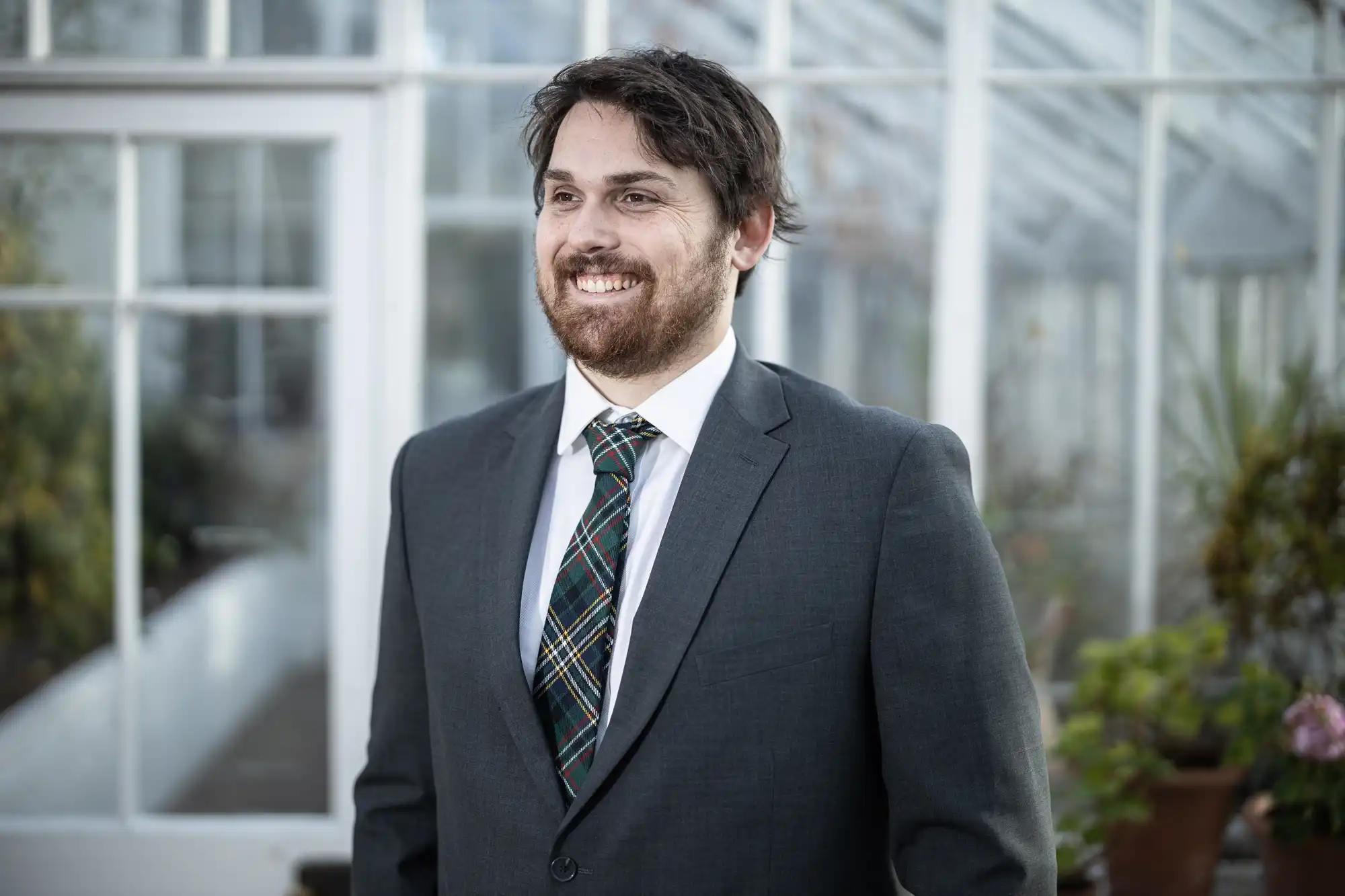 A man with dark hair and a beard, wearing a dark grey suit, white shirt, and checked tie, stands smiling in a bright glasshouse with glass walls and potted plants in the background.
