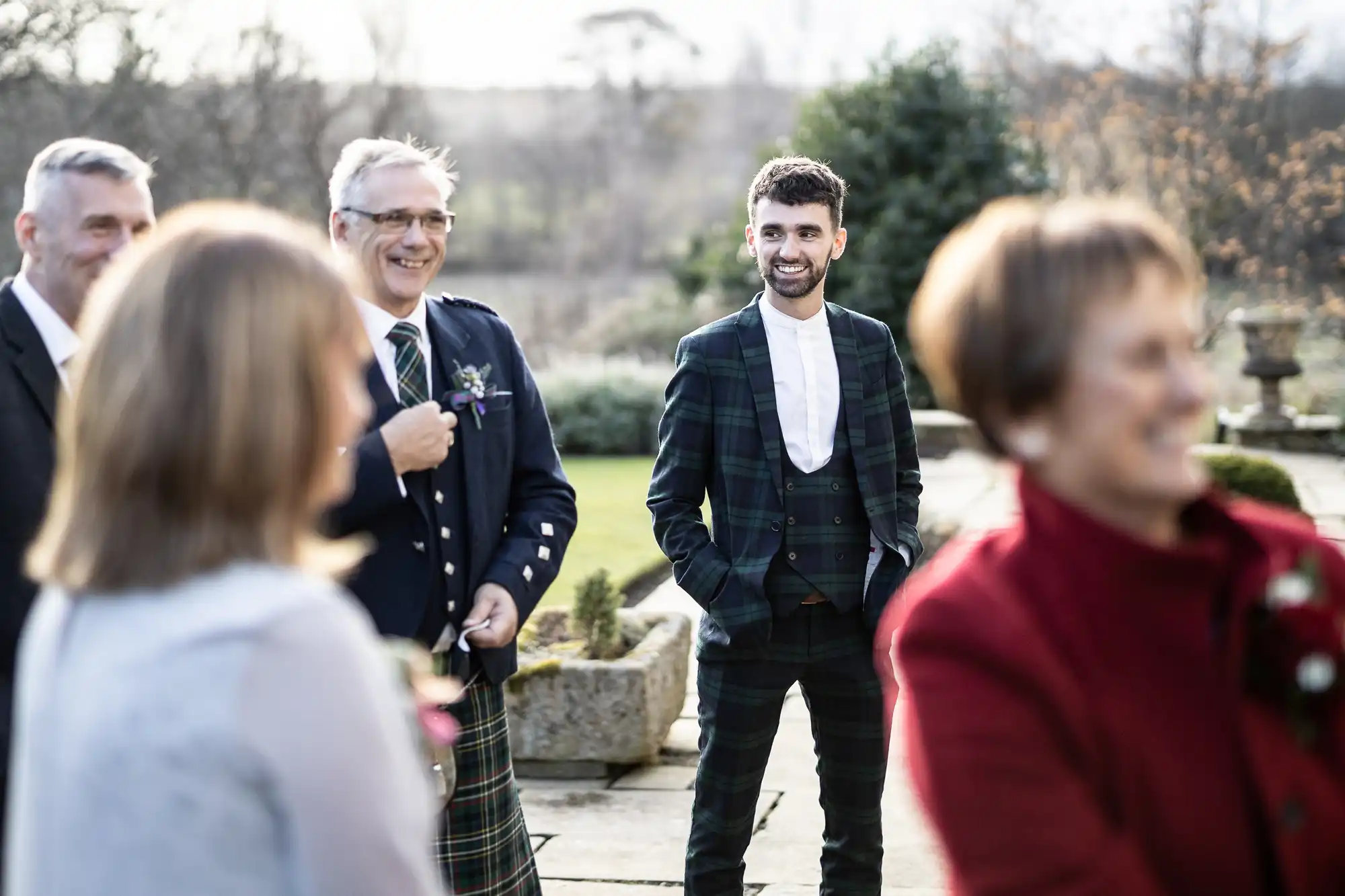 A smiling man in a dark checked suit stands outdoors among four other people, who are slightly out of focus. It appears to be a social gathering or celebration in a garden with stone paths, greenery, and soft natural light.