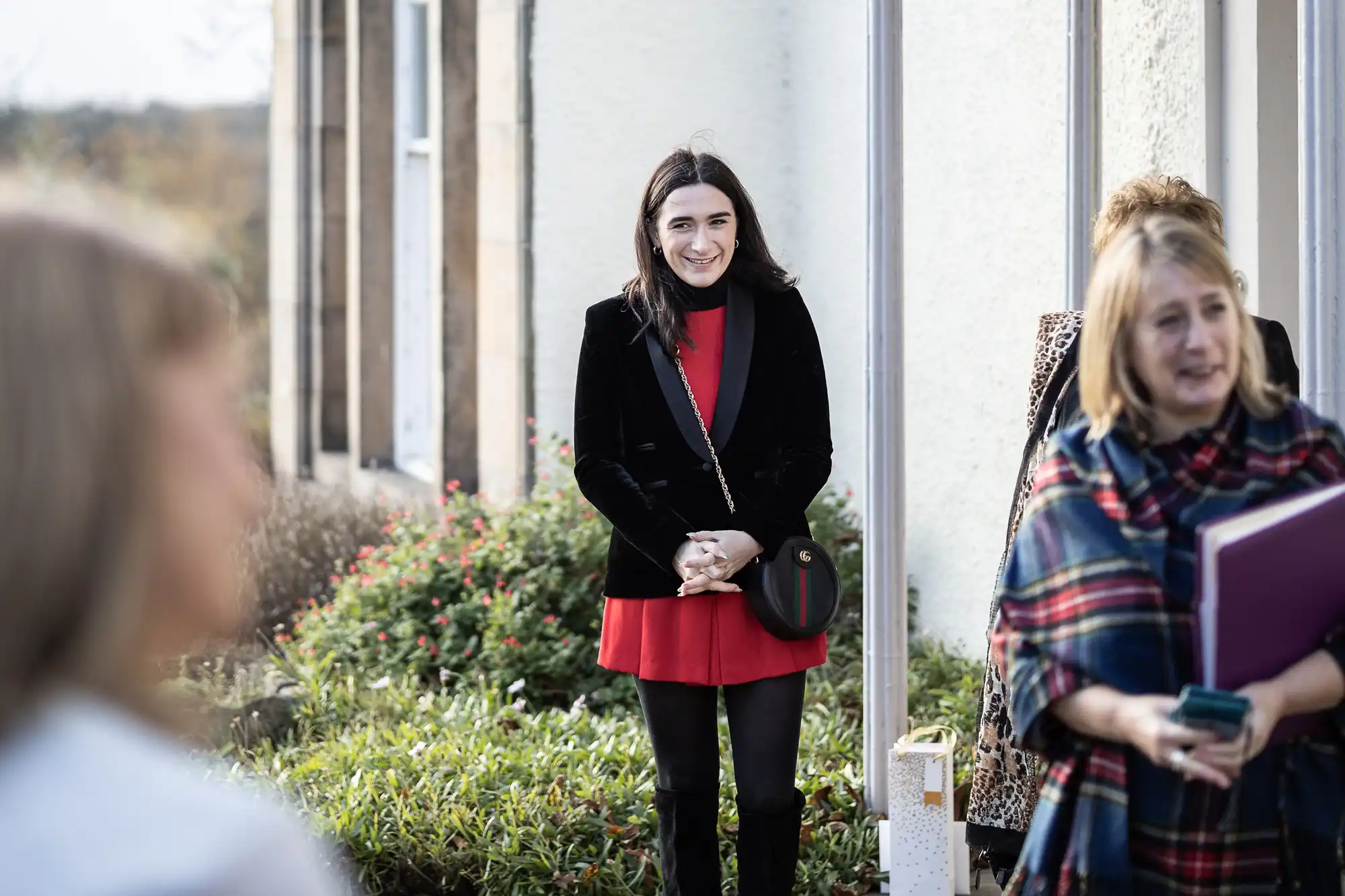 A woman in a black jacket and red dress stands smiling outside by a building, hands clasped. Other women, one holding a purple folder and wearing a checked shawl, stand nearby. Green bushes and flowers line the path. The scene appears casual and social.