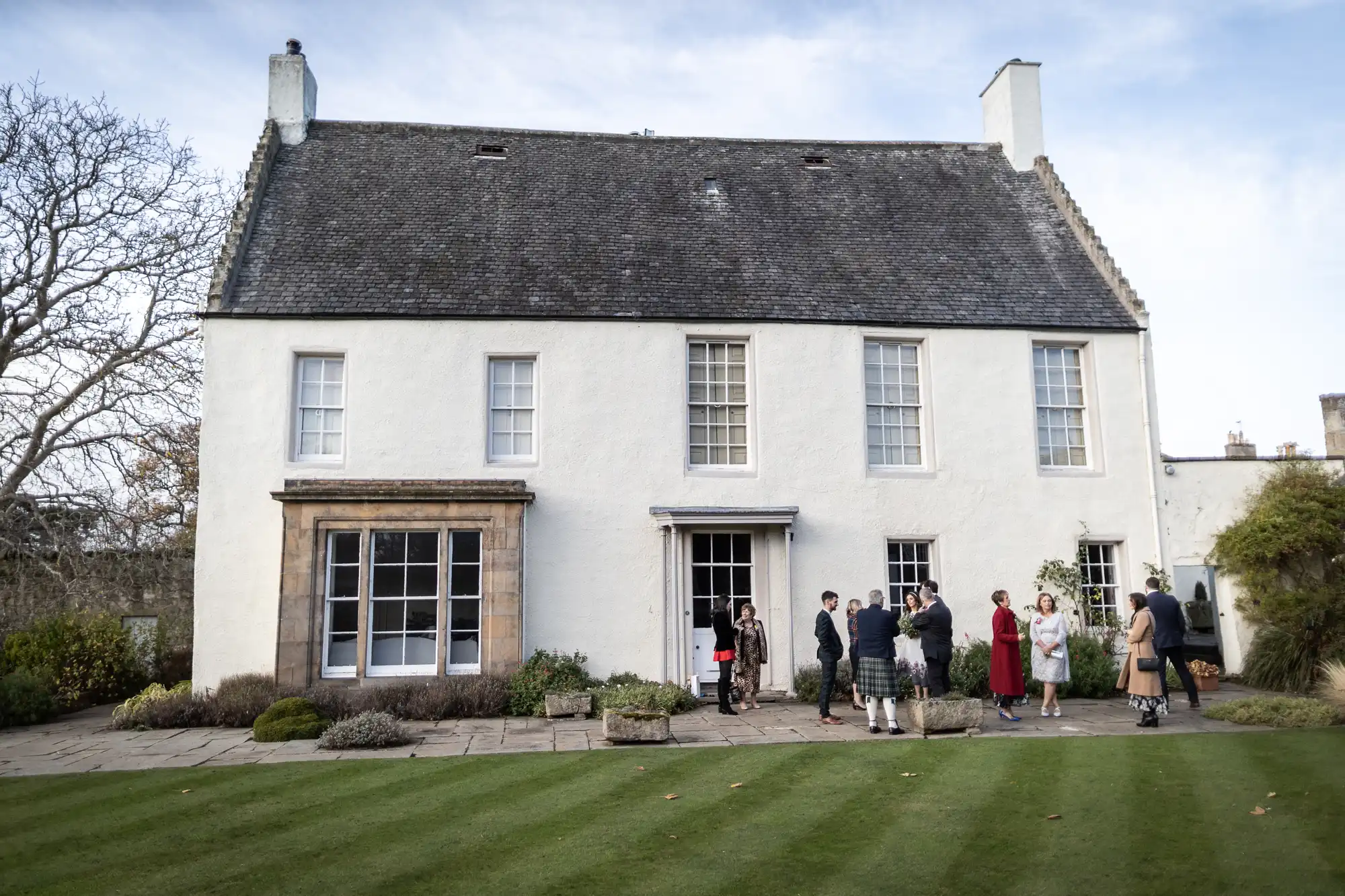 A group of people in formal attire gather outside a large two-storey white house with a steep slate roof and tall windows. The house has a manicured lawn in front and some bare trees and bushes around its stone-paved entrance.
