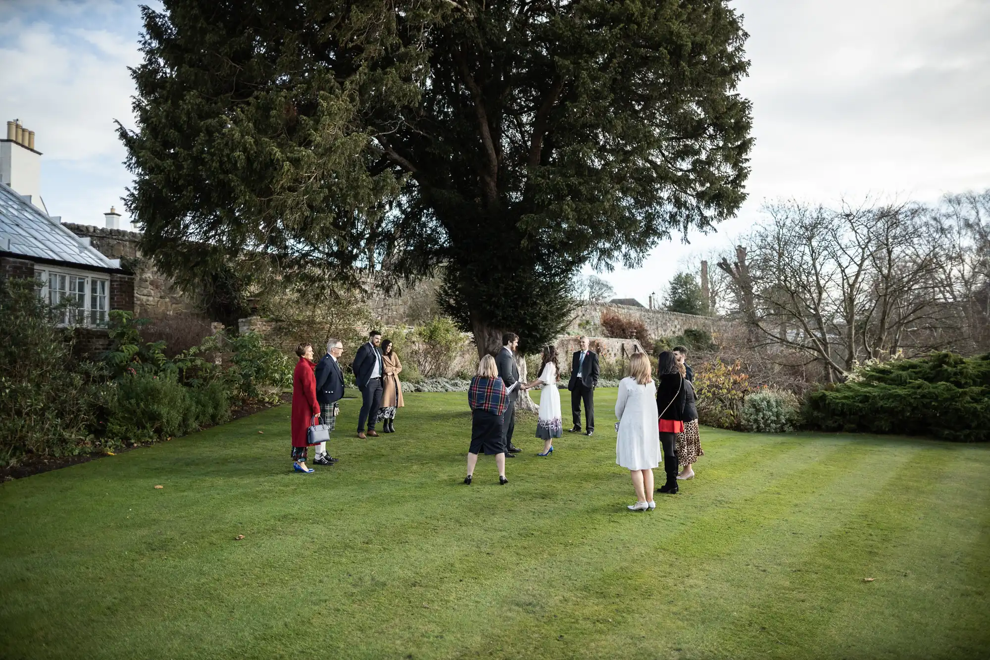 A small group of people, some in kilts and others in dresses and coats, stand in a circle on green grass beneath a large tree, in a garden or park with shrubs and buildings in the background on a cloudy day.