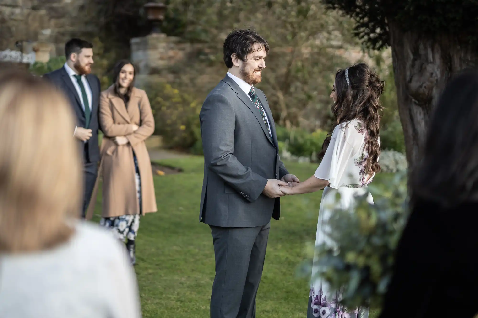 A couple stands facing each other, holding hands and smiling during an outdoor wedding ceremony. The groom wears a grey suit and tie; the bride wears a white dress with floral designs. Guests in coats stand in the background on a grassy lawn with stone walls.