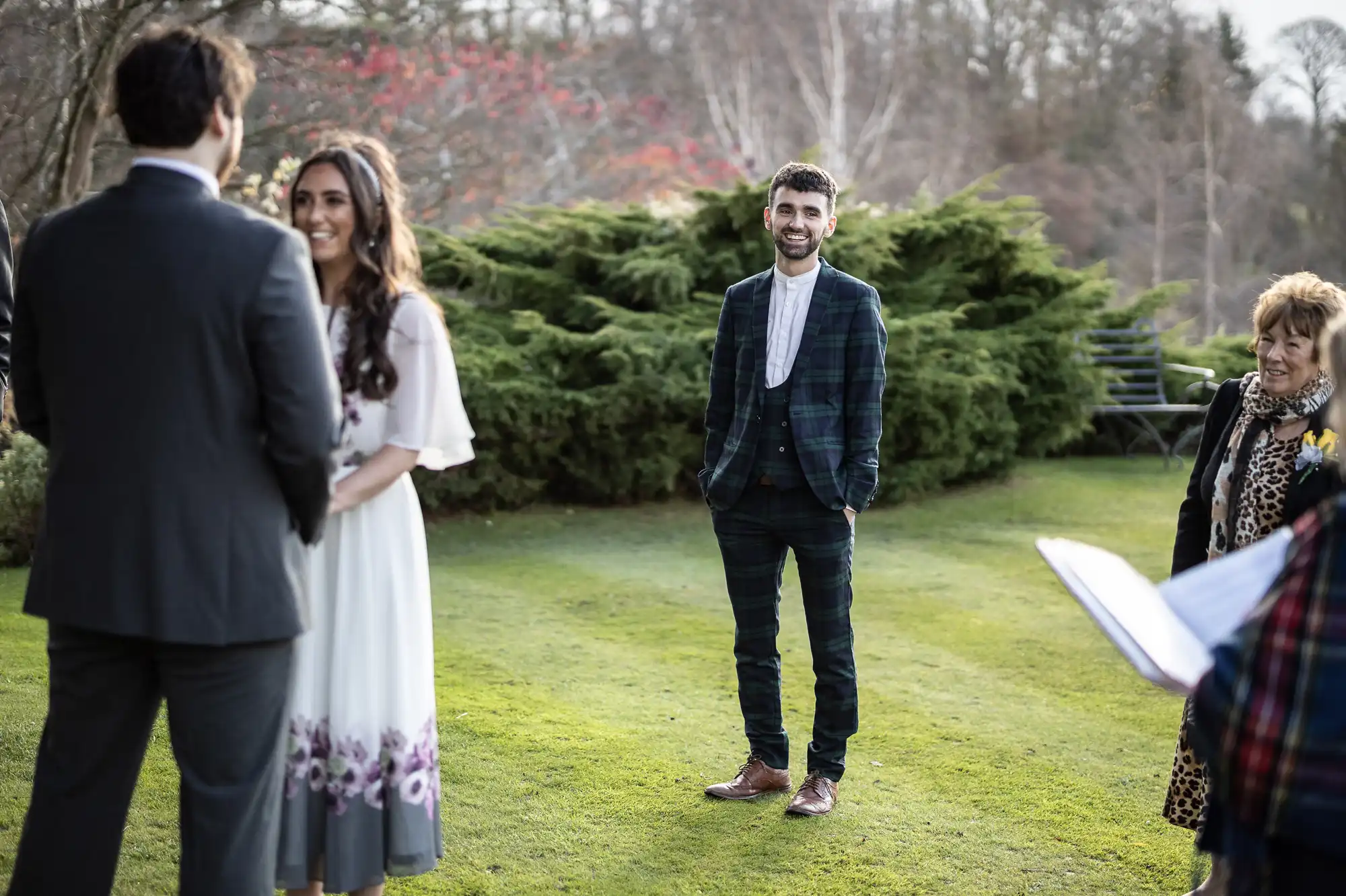 A smiling man in a checked suit stands on a grassy lawn, facing a couple holding hands during an outdoor ceremony. The woman wears a white dress with purple flowers. Another woman on the right holds papers. Bushes and bare trees are in the background.