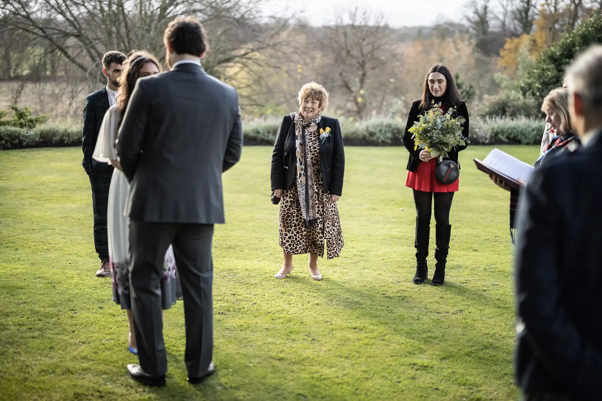 A small outdoor wedding ceremony on a lawn. A couple stands facing each other, a celebrant holds a book, a woman in a leopard print dress smiles, and another woman in a red dress holds a bouquet. Trees and bushes are in the background.