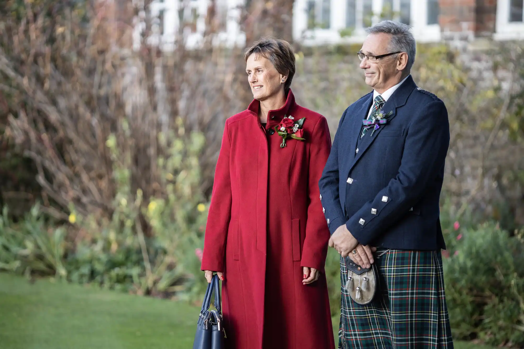 A woman in a red coat holds a blue handbag and stands beside a man in a blue jacket and green tartan kilt, both wearing buttonholes. They are outdoors on grass, with blurred plants and a brick building with windows in the background.