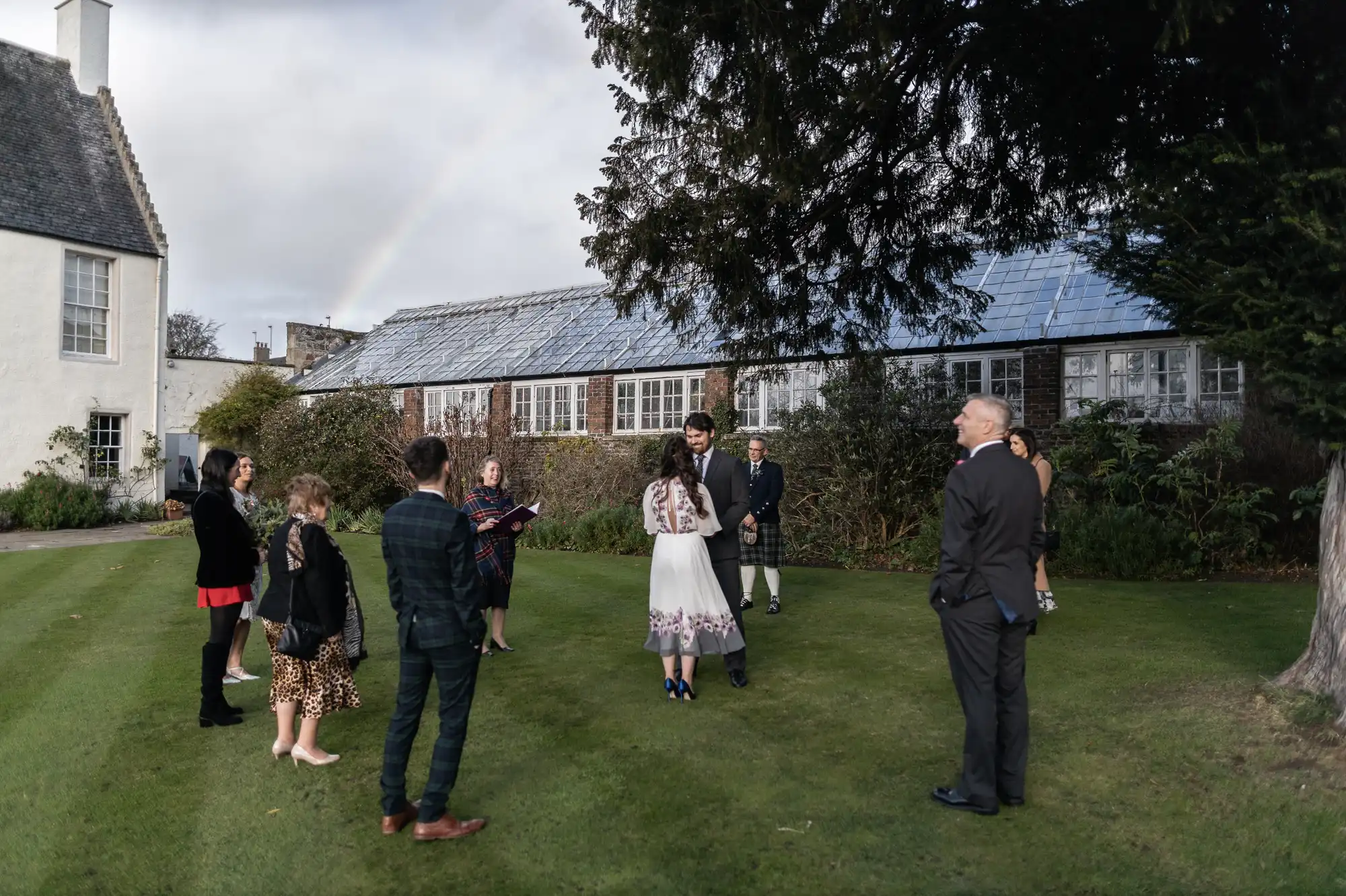 A small outdoor wedding ceremony with ten people standing spaced in a circle on a lawn. The bride and groom face each other in the centre. Guests wear semi-formal clothes; some are in dresses, some in suits. Trees, buildings, and a faint rainbow appear in the background.