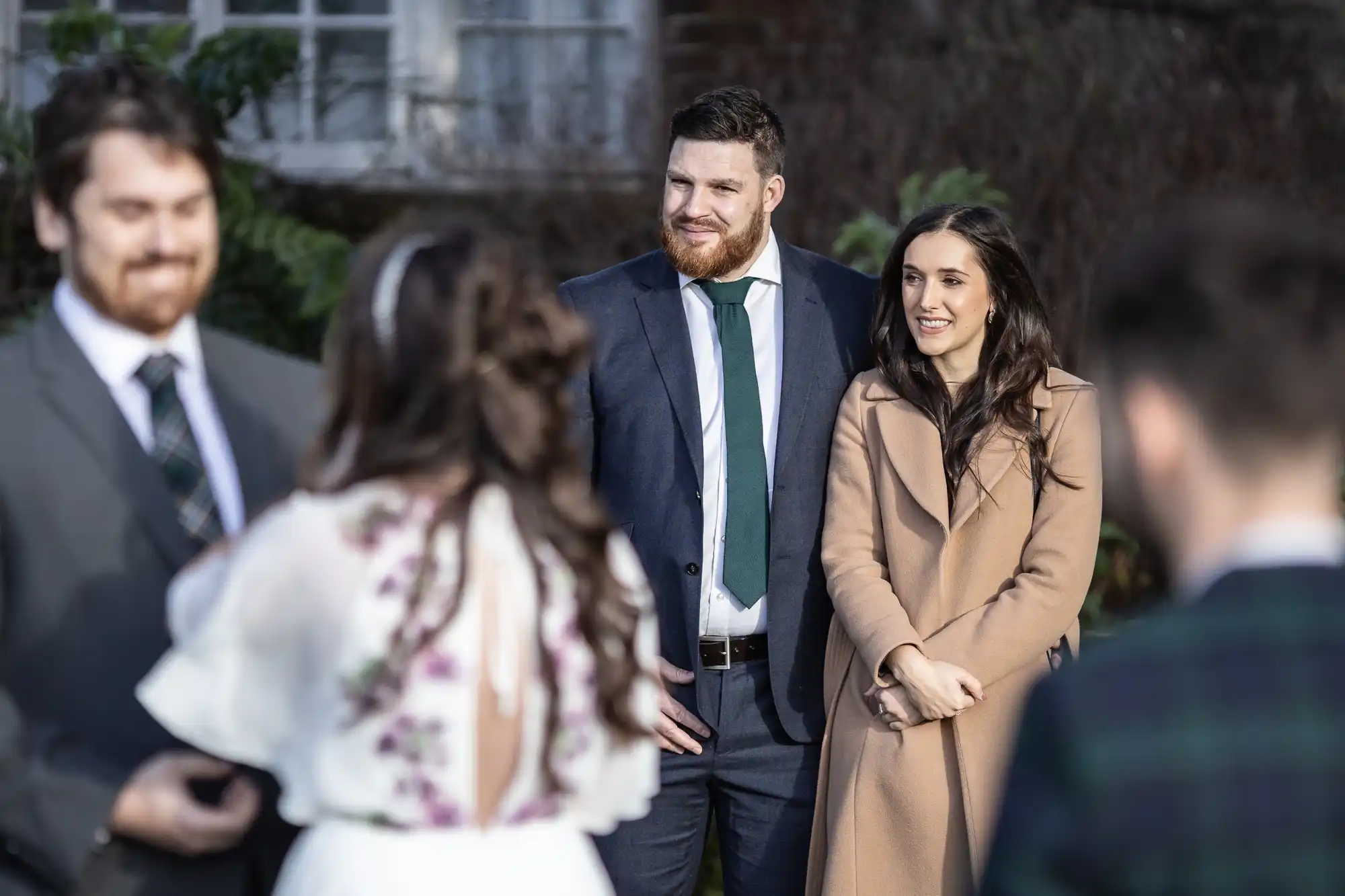 A man in a dark suit and green tie stands next to a woman in a tan coat. They are smiling outdoors among a group of people, including a person in a white dress with floral details. The mood is cheerful, suggesting a social or celebratory event.