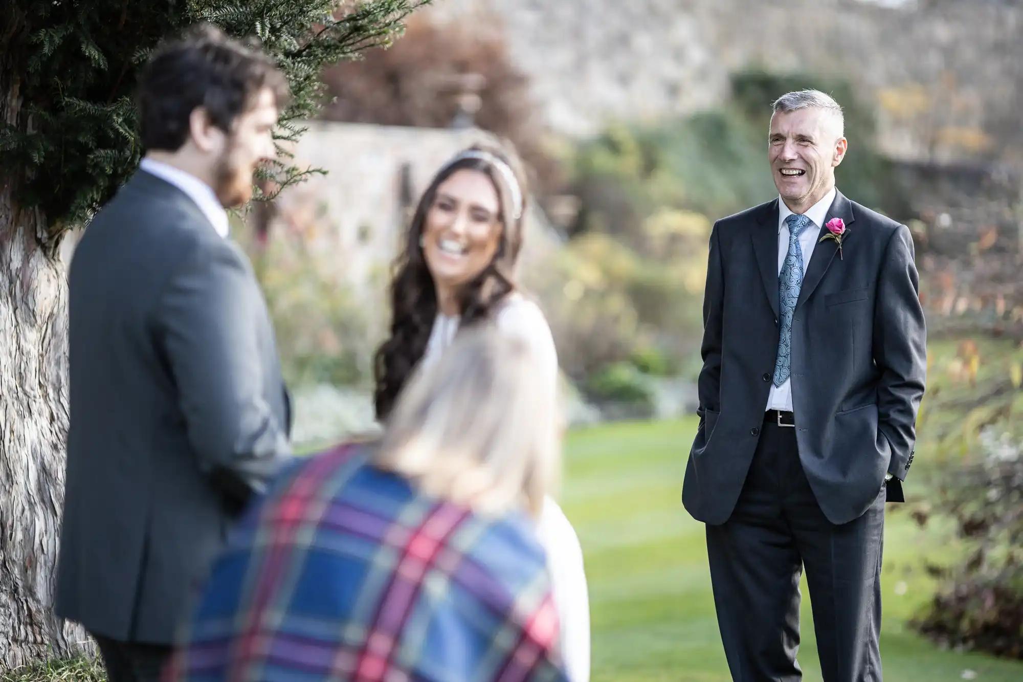 Three people stand outdoors, smiling and laughing. A man in a suit with a pink buttonhole stands on the right. In the foreground, a woman with long hair faces away, and a man and woman, both dressed up, are smiling and interacting, with trees and greenery in the background.
