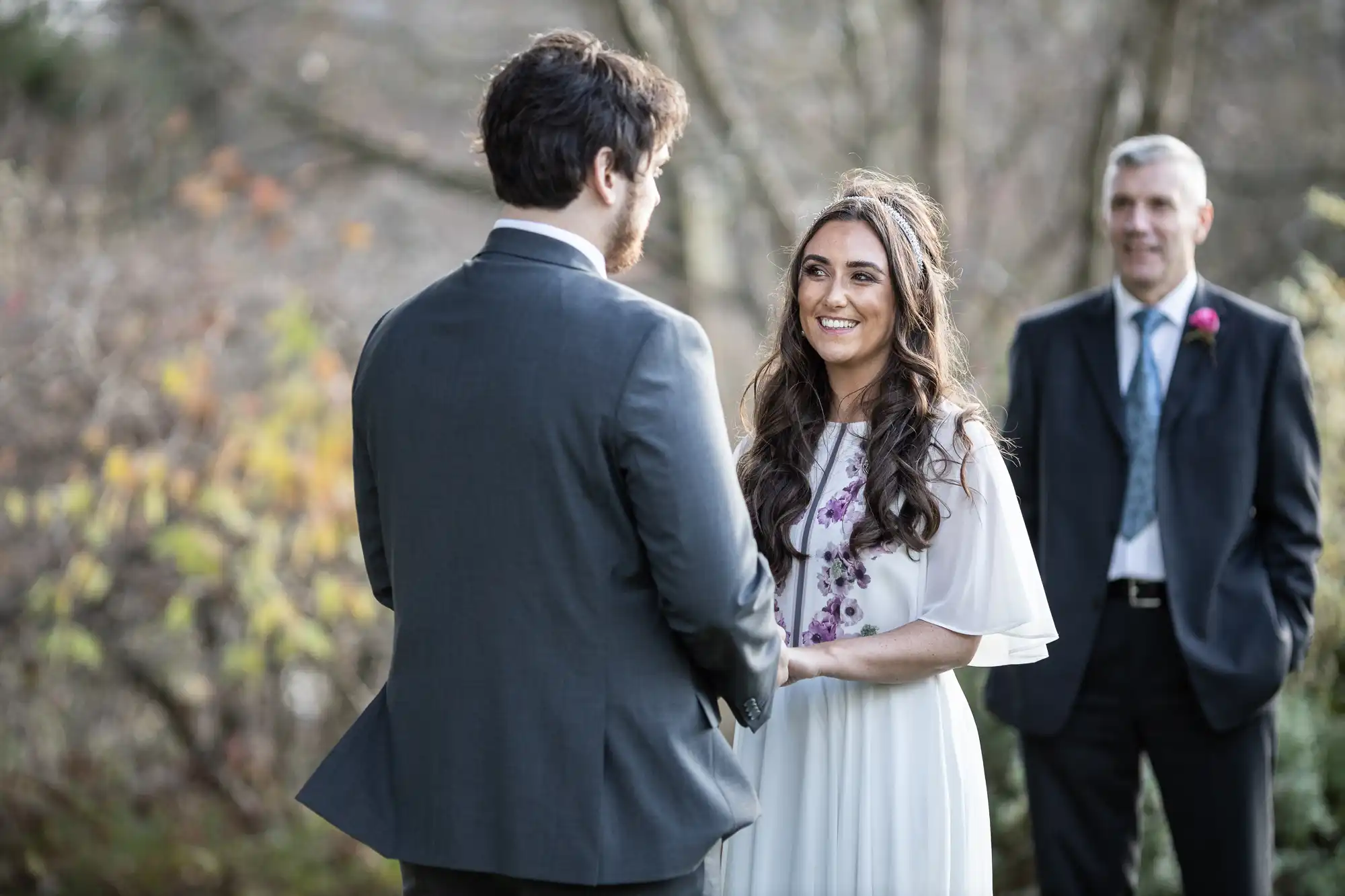 A bride in a white dress with long brown hair smiles at a groom in a grey suit during an outdoor wedding ceremony. A man in a dark suit stands behind them, watching. The background features blurred trees and soft natural light.