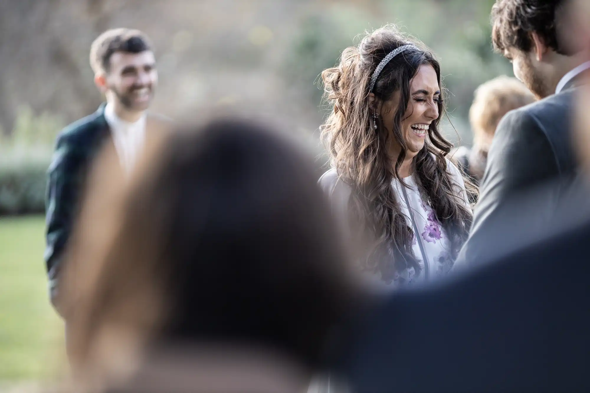 A woman with long wavy brown hair, smiling brightly, stands outdoors at a wedding ceremony. She faces a man in a suit. The background is blurred, with another man in focus smiling, and several indistinct people in the foreground.