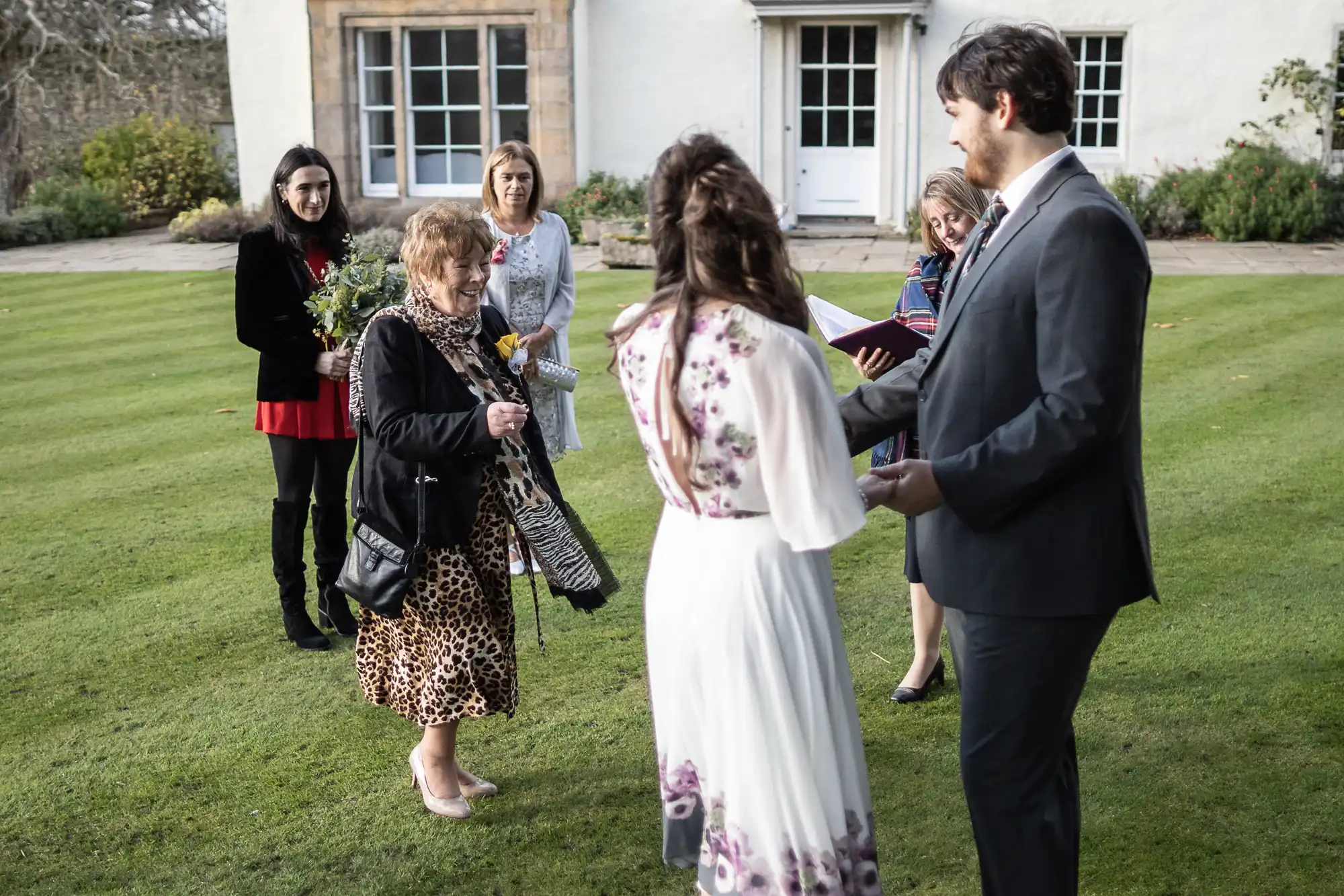 A small outdoor wedding ceremony on a lawn features a bride in a white dress with purple accents and a groom in a grey suit, facing each other. Four women stand nearby; one holds flowers, others look on, with a house in the background.
