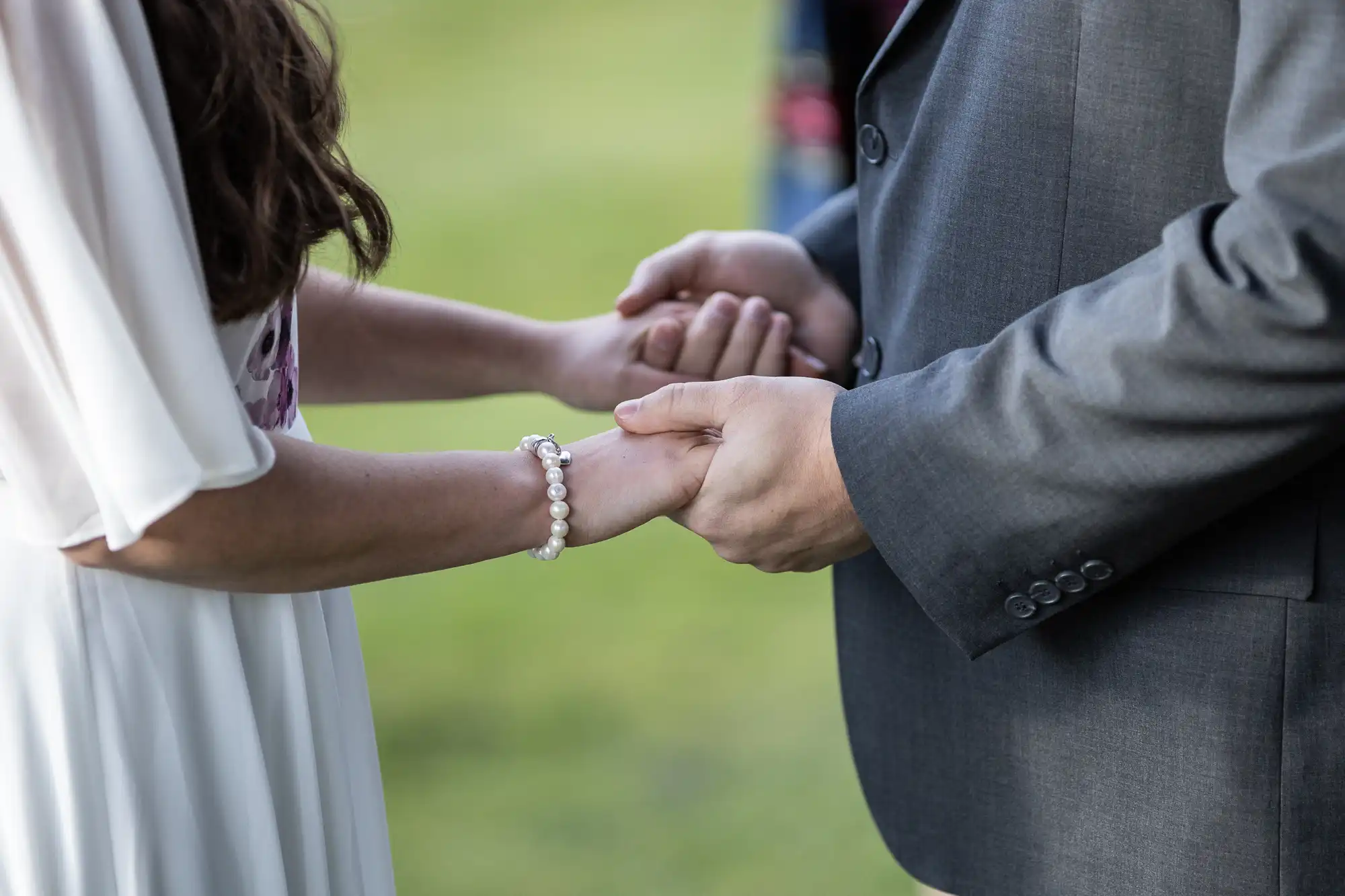 A close-up of two people holding hands outdoors. One person wears a white dress and a pearl bracelet, while the other wears a grey suit. Only their torsos and hands are visible, with a blurred green background behind them.