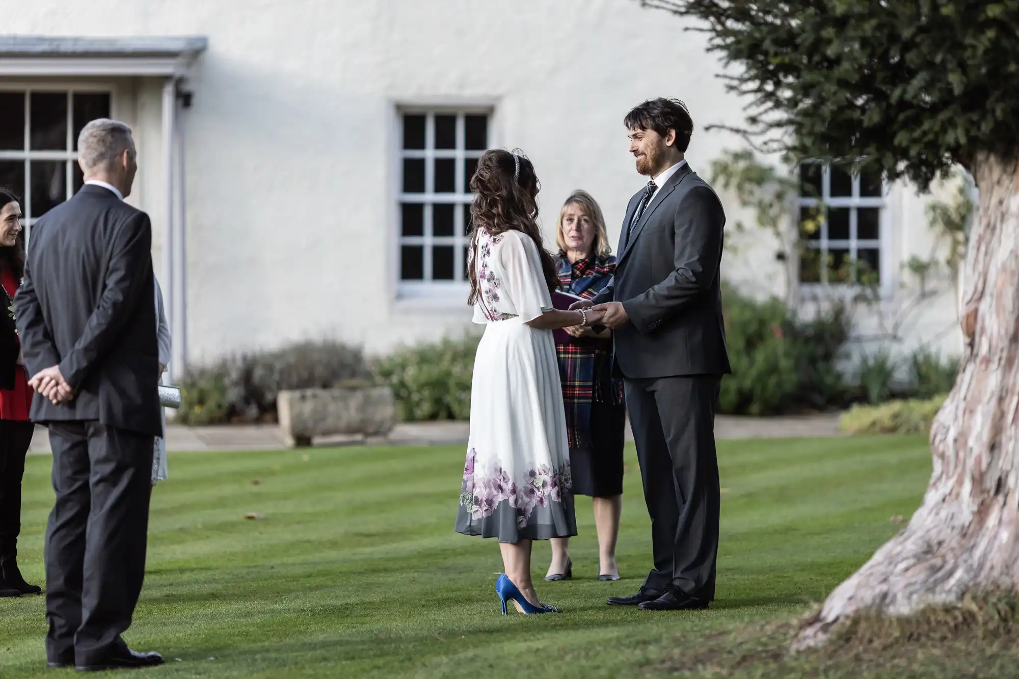 A couple stands facing each other, holding hands, during an outdoor wedding ceremony on grass. The bride wears a white dress with purple flowers and blue heels; the groom is in a dark suit. Guests and an officiant stand nearby in front of a white building with windows.