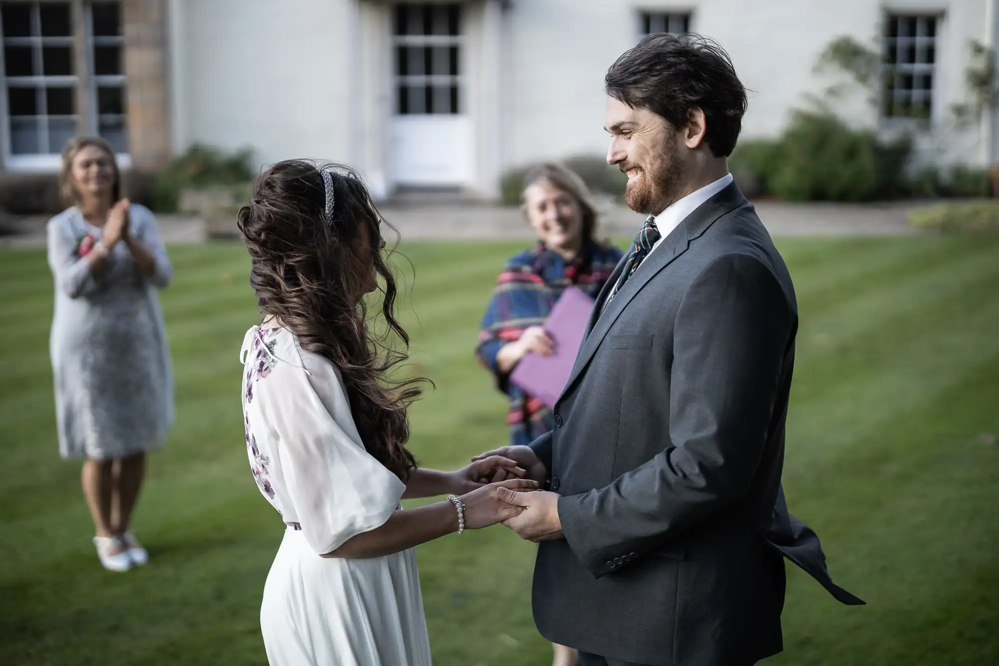 A couple stands facing each other outside, holding hands and smiling, dressed in formal attire. A woman in a dress claps in the background whilst another person stands behind them, holding a folder and smiling, suggesting a small wedding ceremony on a lawn.
