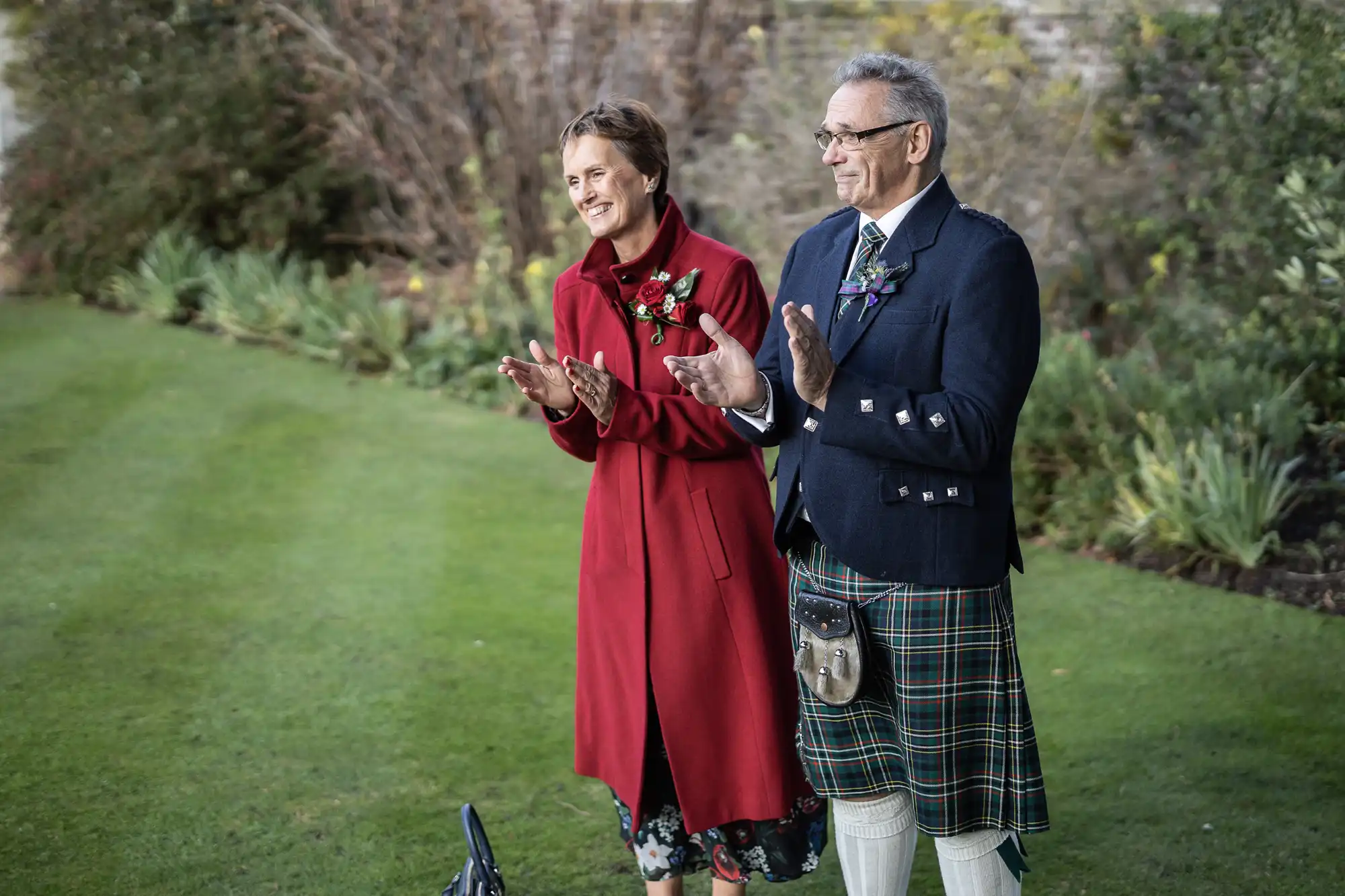 A woman in a long red coat and a man in a blue jacket, kilt, and sporran stand on grass, smiling and clapping. They appear to be outdoors in a garden with green plants and shrubs in the background. Both look happy and engaged.