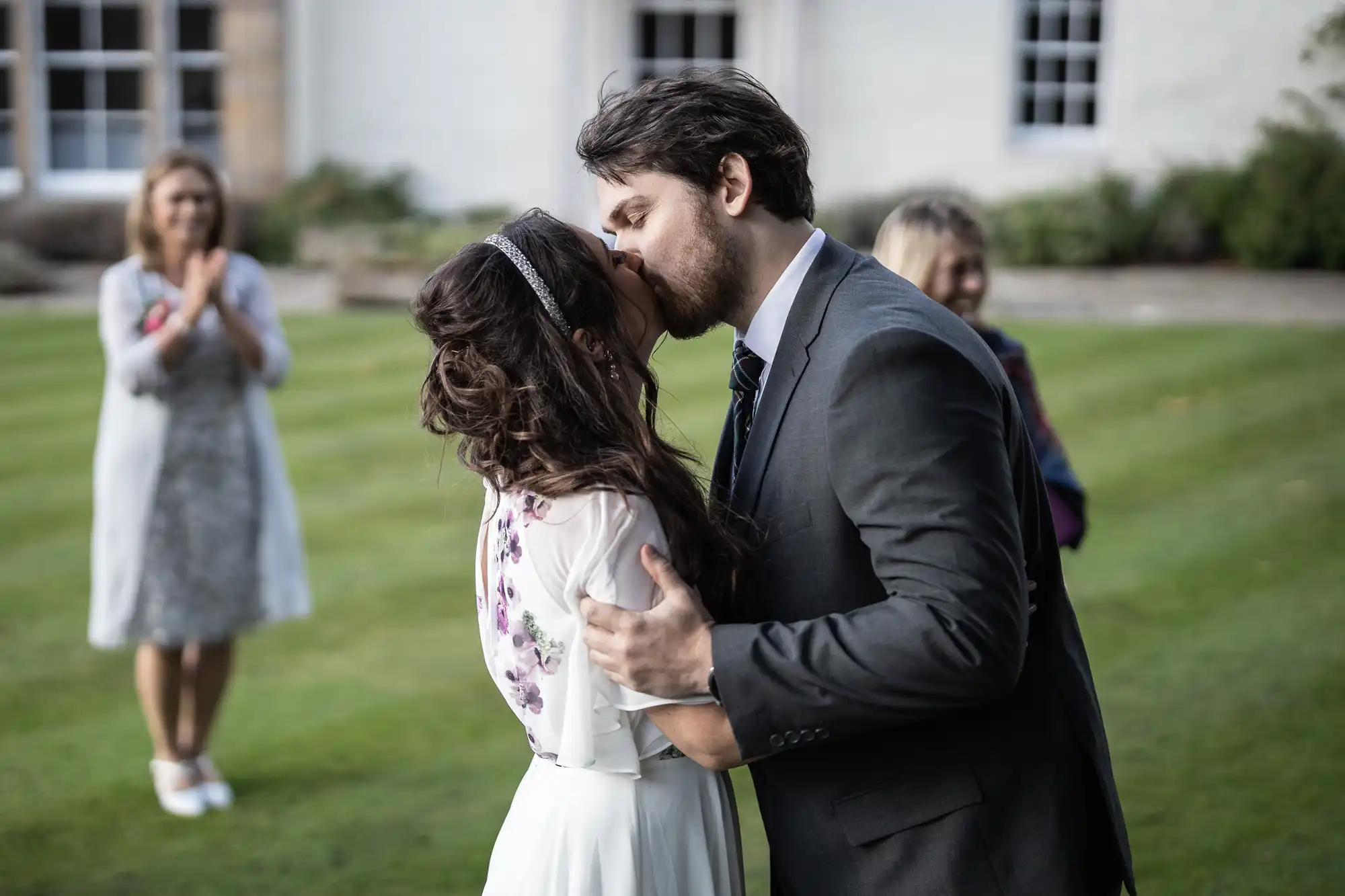 A bride and groom share a kiss outdoors on green grass. The bride wears a white dress with floral details; the groom is in a dark suit. In the background, two women in dresses watch and clap, with a blurred white building visible.
