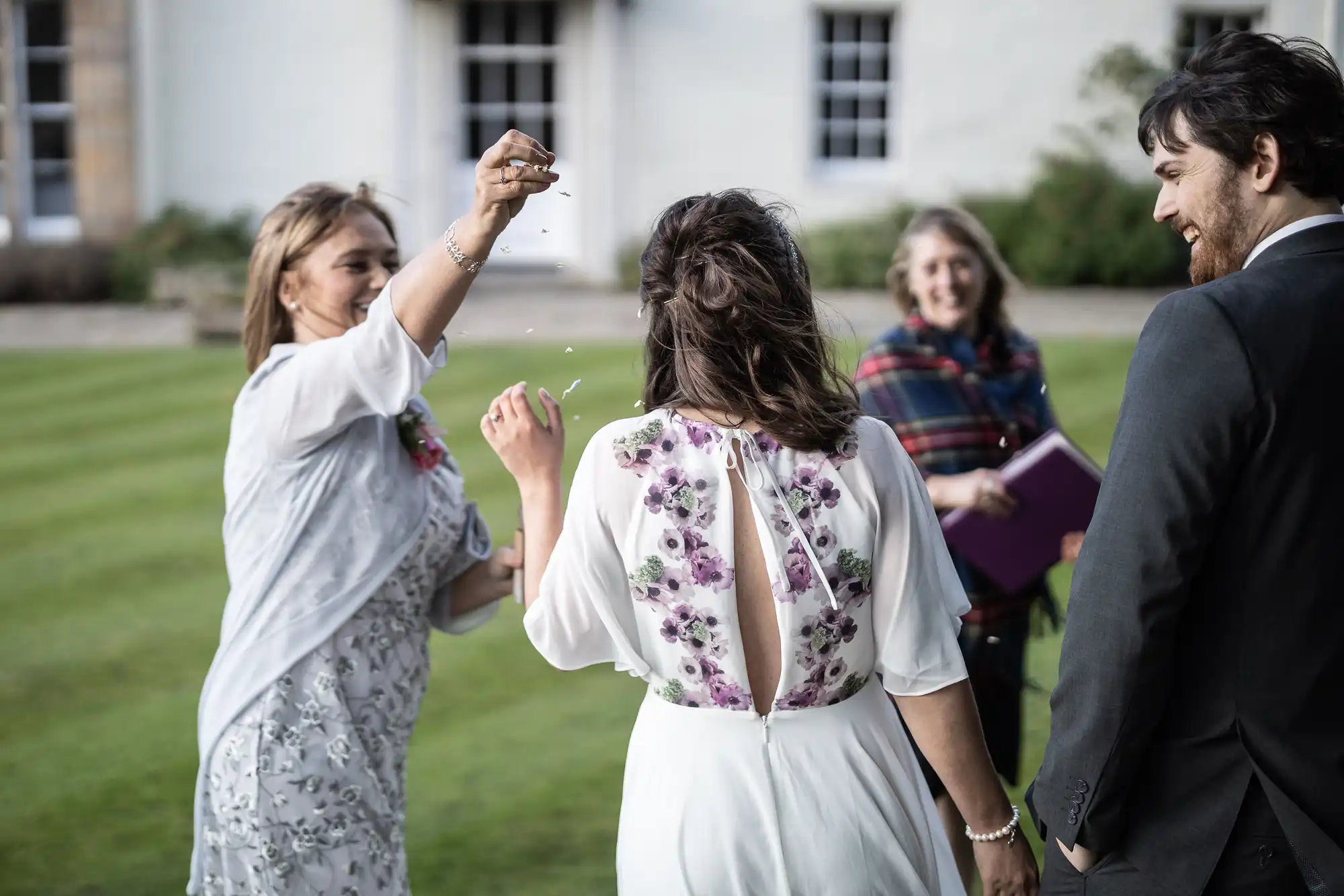 A woman in a white floral dress sprinkles confetti over another woman in a white dress with purple floral details. Two people, one holding a book and another in a dark suit, smile and watch outside on green grass near a large building.