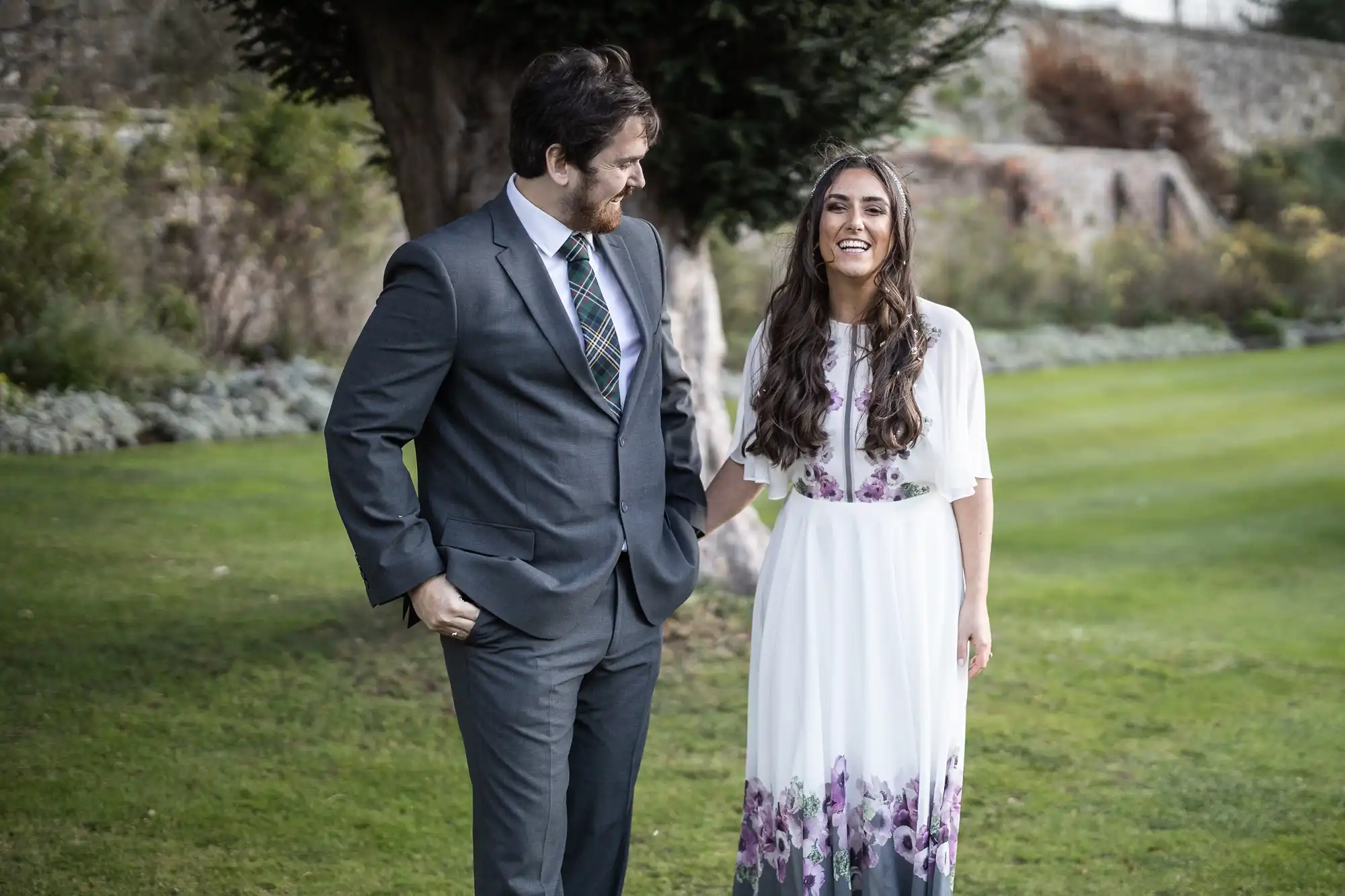 A man in a grey suit and checked tie stands next to a smiling woman in a white dress with purple floral patterns. They are outdoors on green grass, with trees and a stone wall in the background, enjoying a moment together.