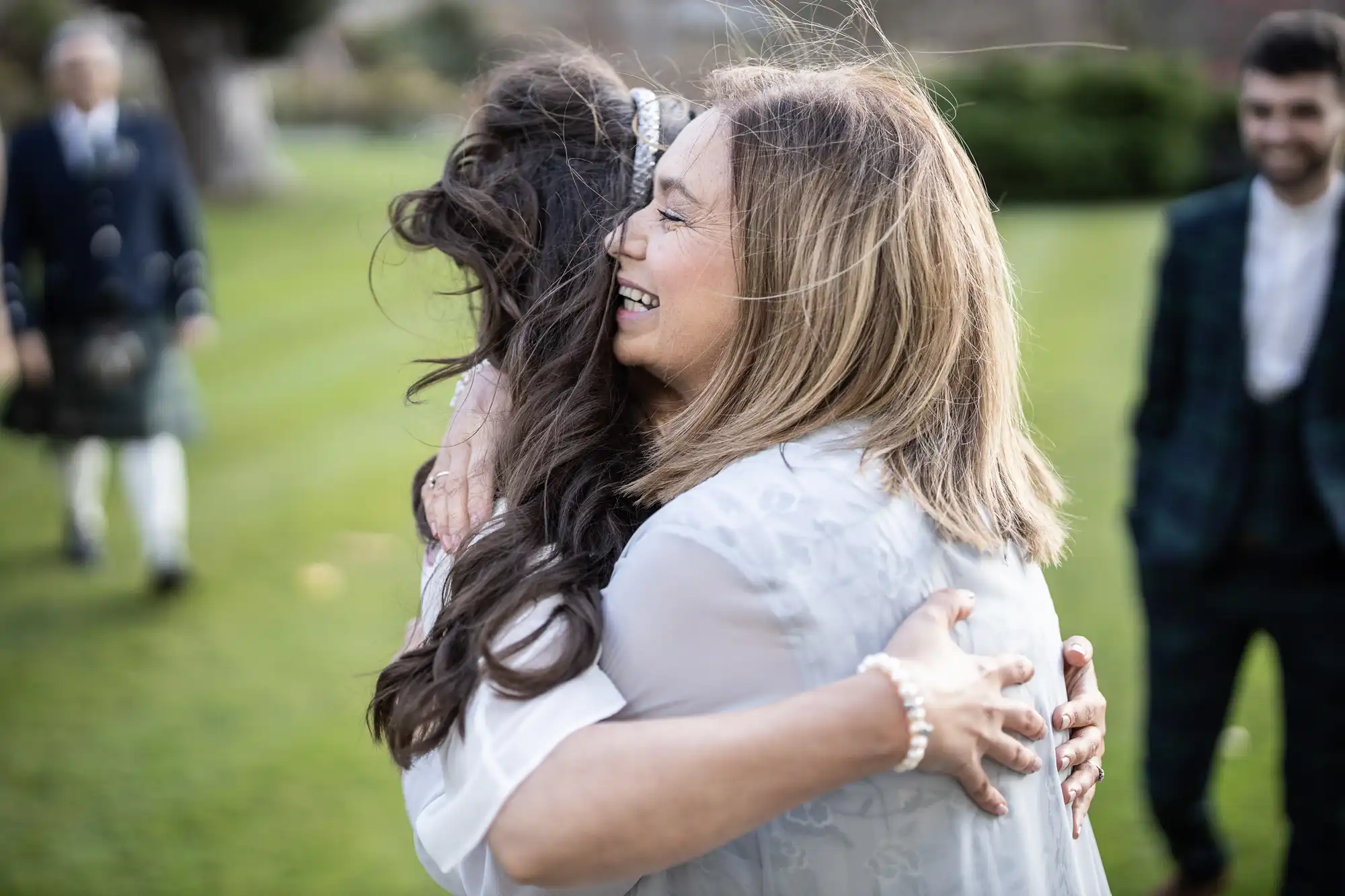 Two women embrace warmly outdoors on a grassy lawn. One woman faces the camera, smiling with eyes closed, while the other’s face is hidden. People in formal attire stand in the blurred background, suggesting a celebratory event.