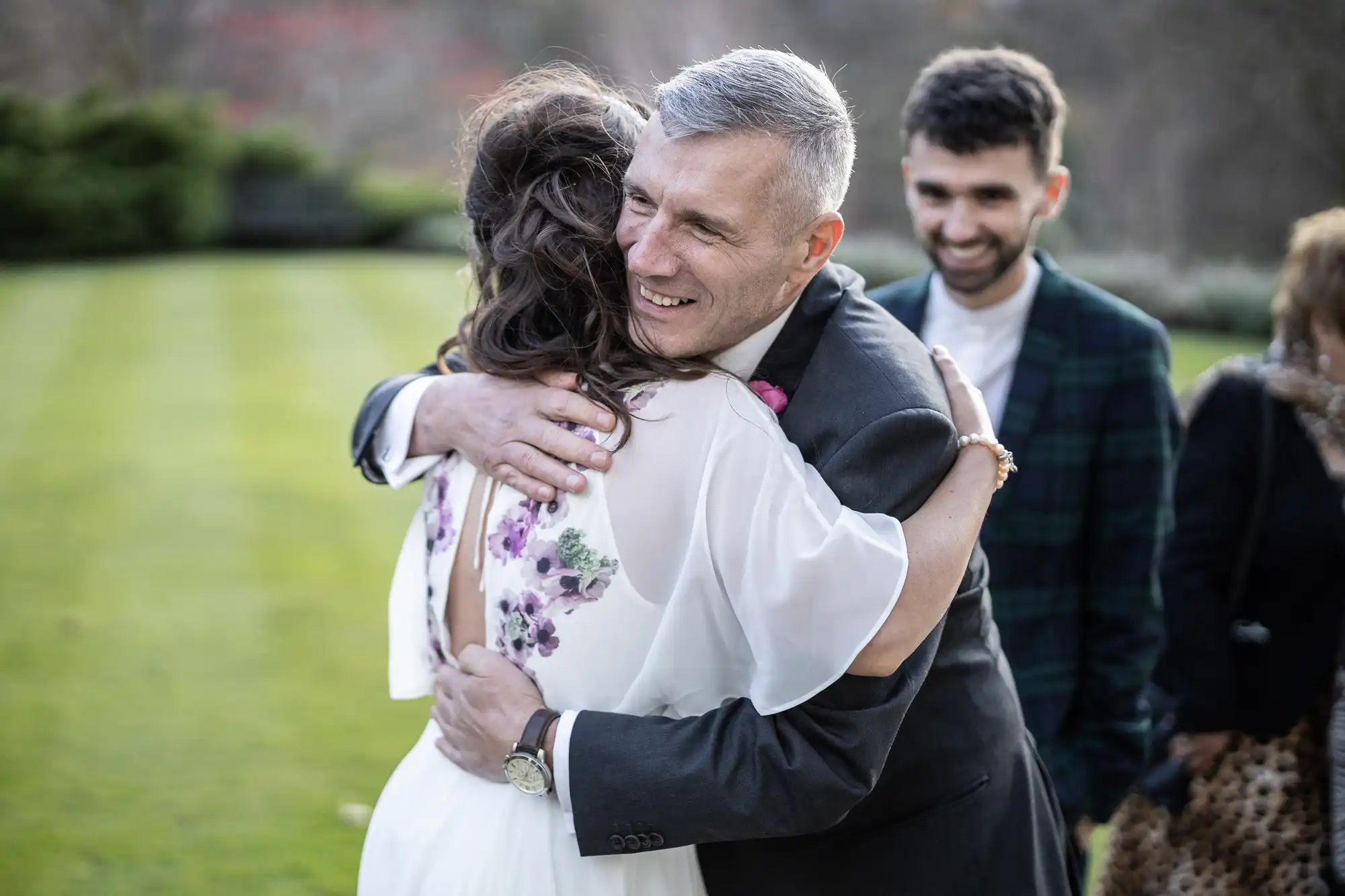 A man in a suit hugs a woman in a white dress with floral details outdoors on a lawn. Both appear joyful. Another man in a dark checked jacket smiles in the background. The scene suggests a celebratory or emotional moment, possibly at a wedding.