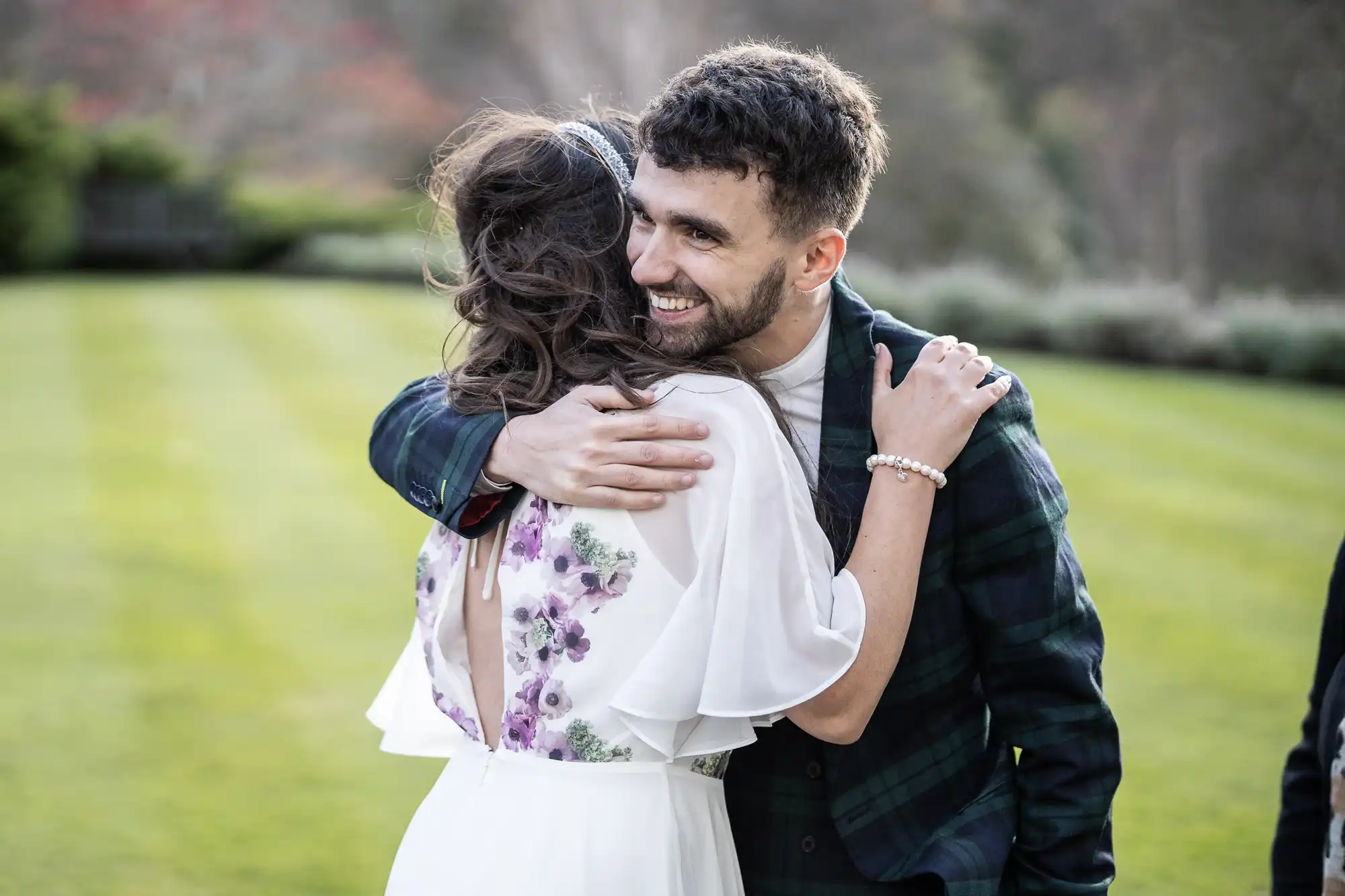 A man with dark hair and a beard smiles as he warmly hugs a woman in a white dress with purple floral patterns. They stand outdoors on a neatly mown lawn, with trees in the background, expressing joy and affection.