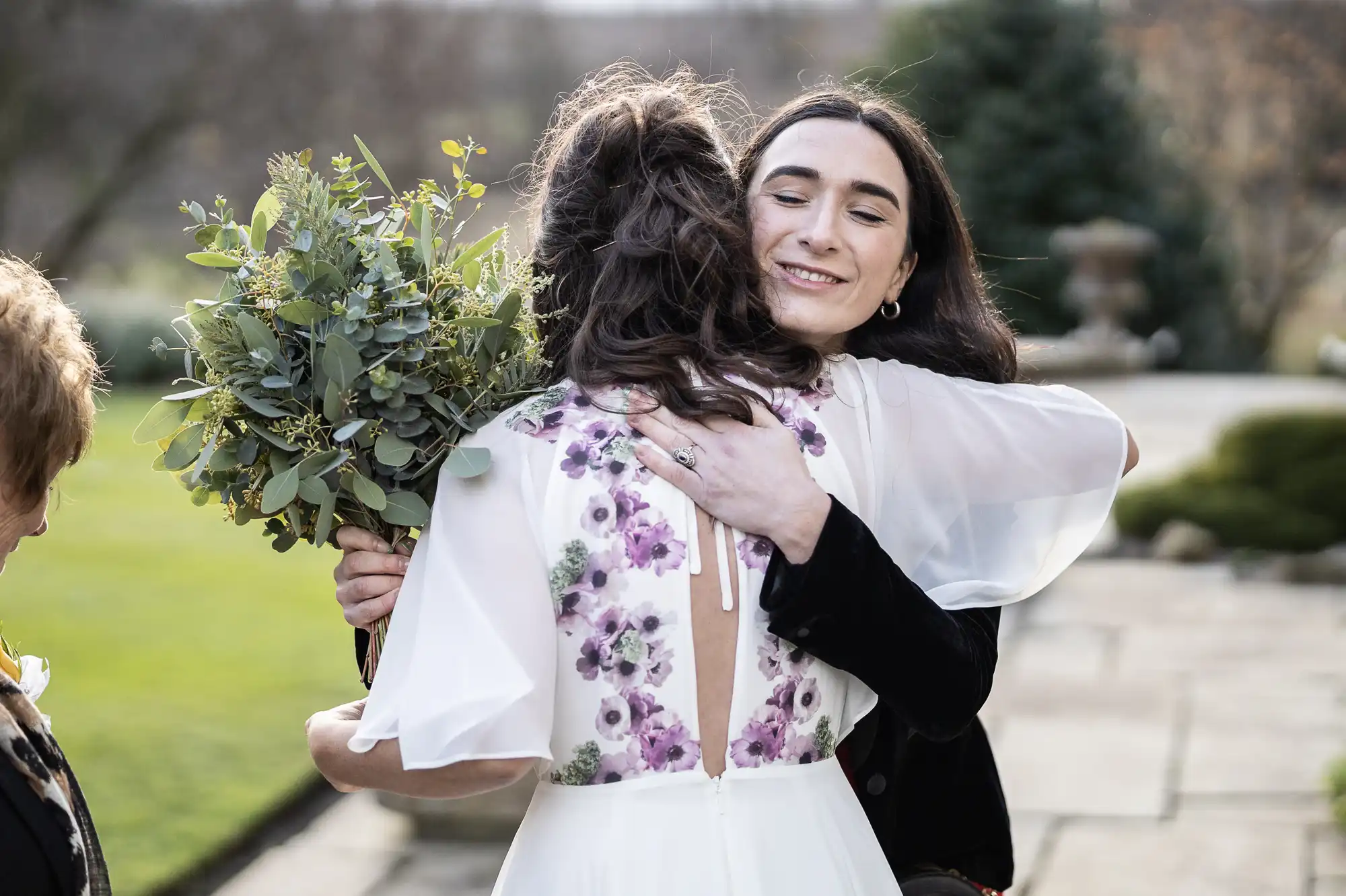 A bride in a white dress with purple floral details hugs a smiling woman outdoors. The bride holds a large bouquet of greenery. The scene is bright, with blurred grass, stone, and trees in the background.