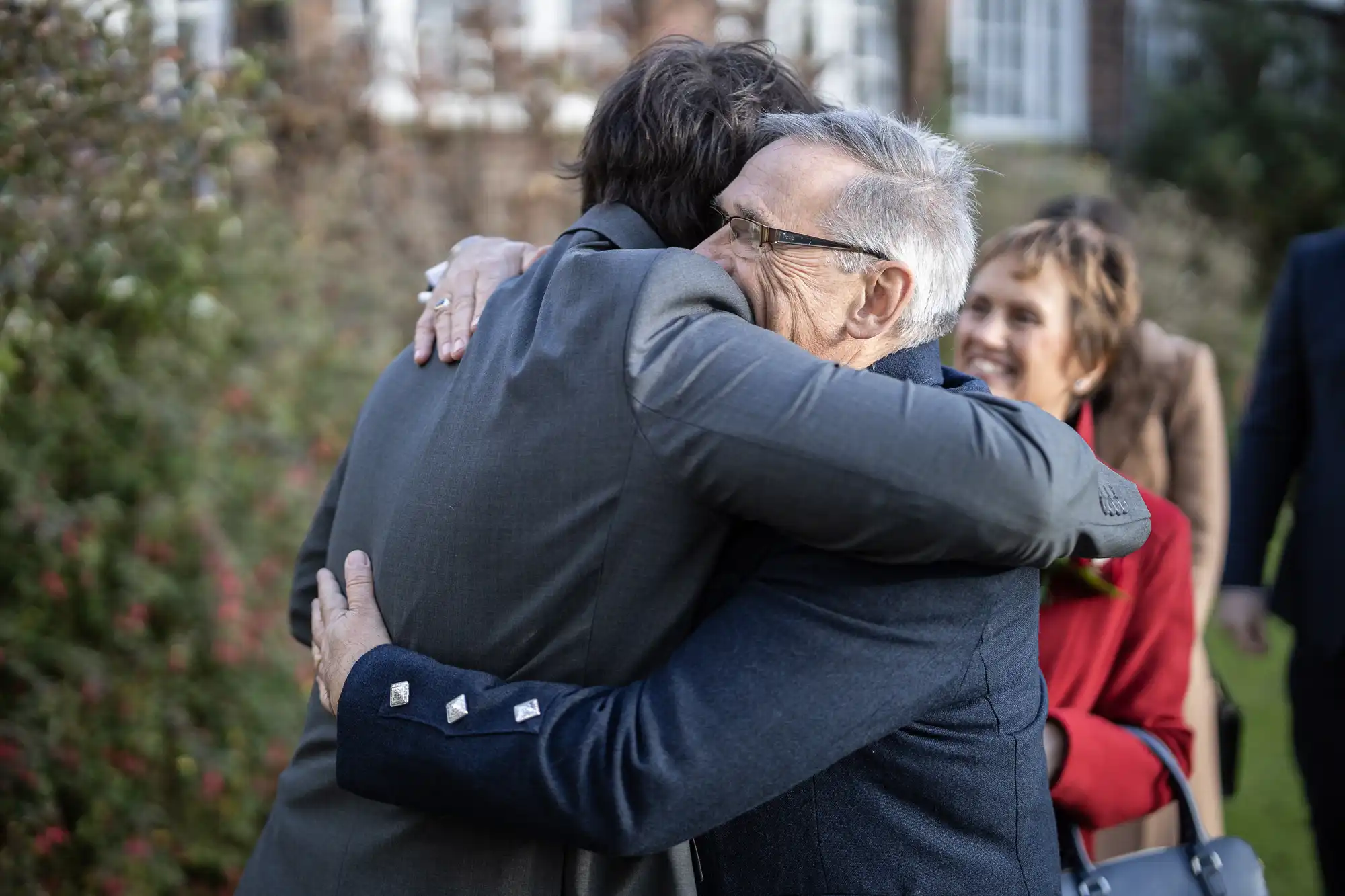 Two men warmly embrace outdoors, one older with grey hair and glasses, the other with brown hair in a grey suit. A smiling woman in a red coat stands nearby, holding a green leafy item. Blurred greenery and building are visible in the background.