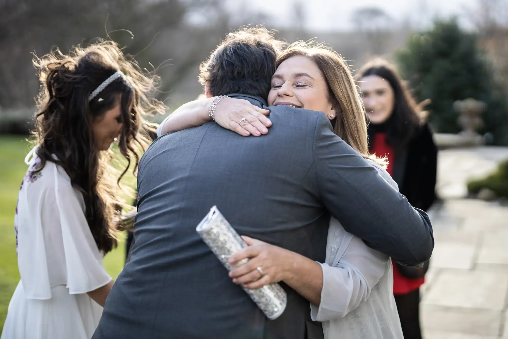 A woman smiles warmly as she hugs a man in a grey suit outdoors. She holds a sparkling silver clutch. Another woman in white and a woman in a red dress stand nearby on a stone path, with greenery and trees in the background.