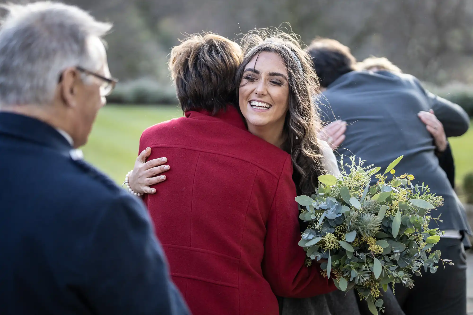 A smiling woman with long hair and a bouquet hugs an older woman in a red coat outdoors; other people in the background also embrace, with greenery and blurred trees in the distance.